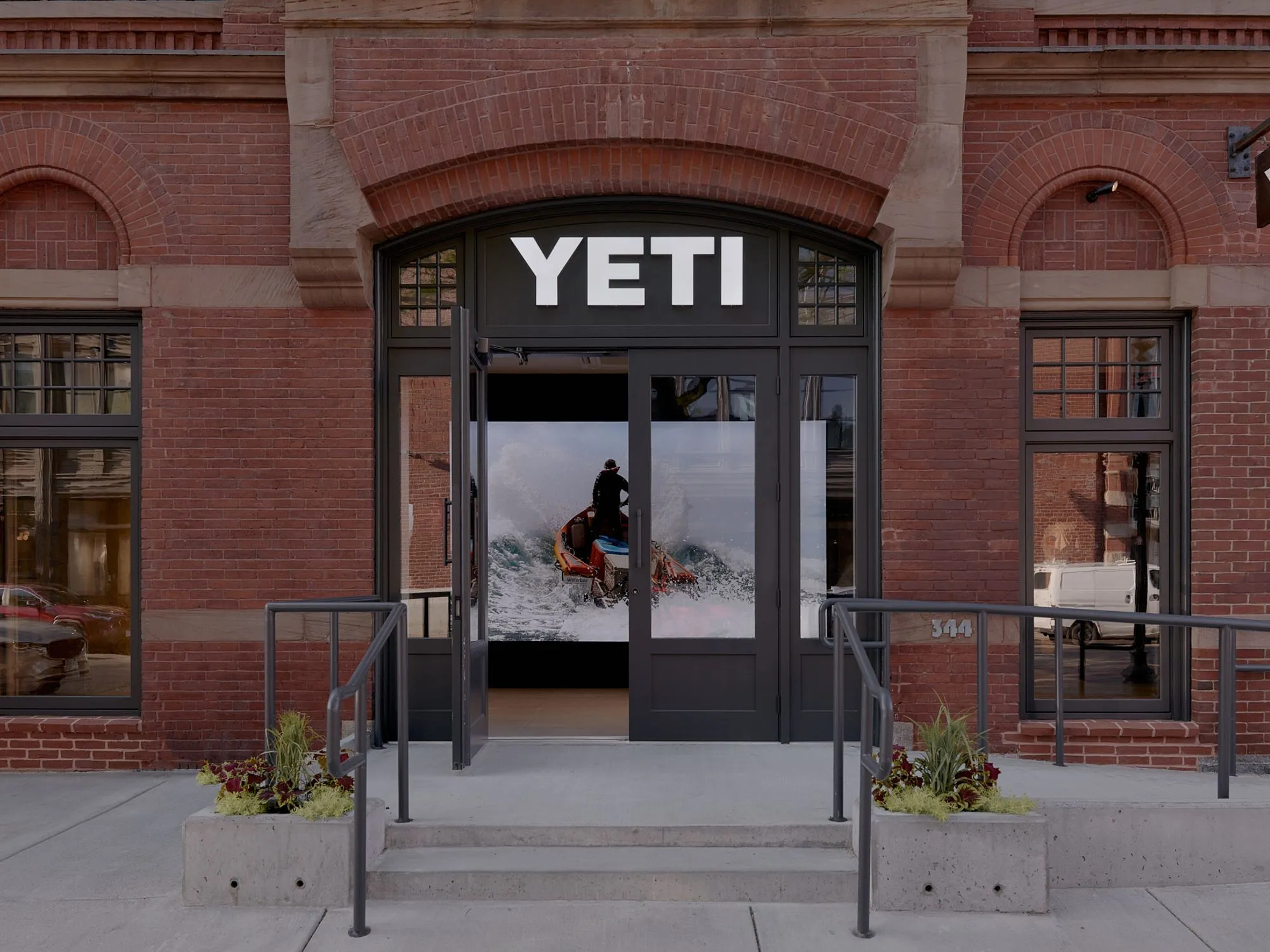 A modern YETI retail storefront features a dark-framed entrance with large glass doors set within a historic red brick building with arched windows and ornamental detailing; the storefront displays an action photograph of a person with outdoor gear against a snowy mountain landscape.
