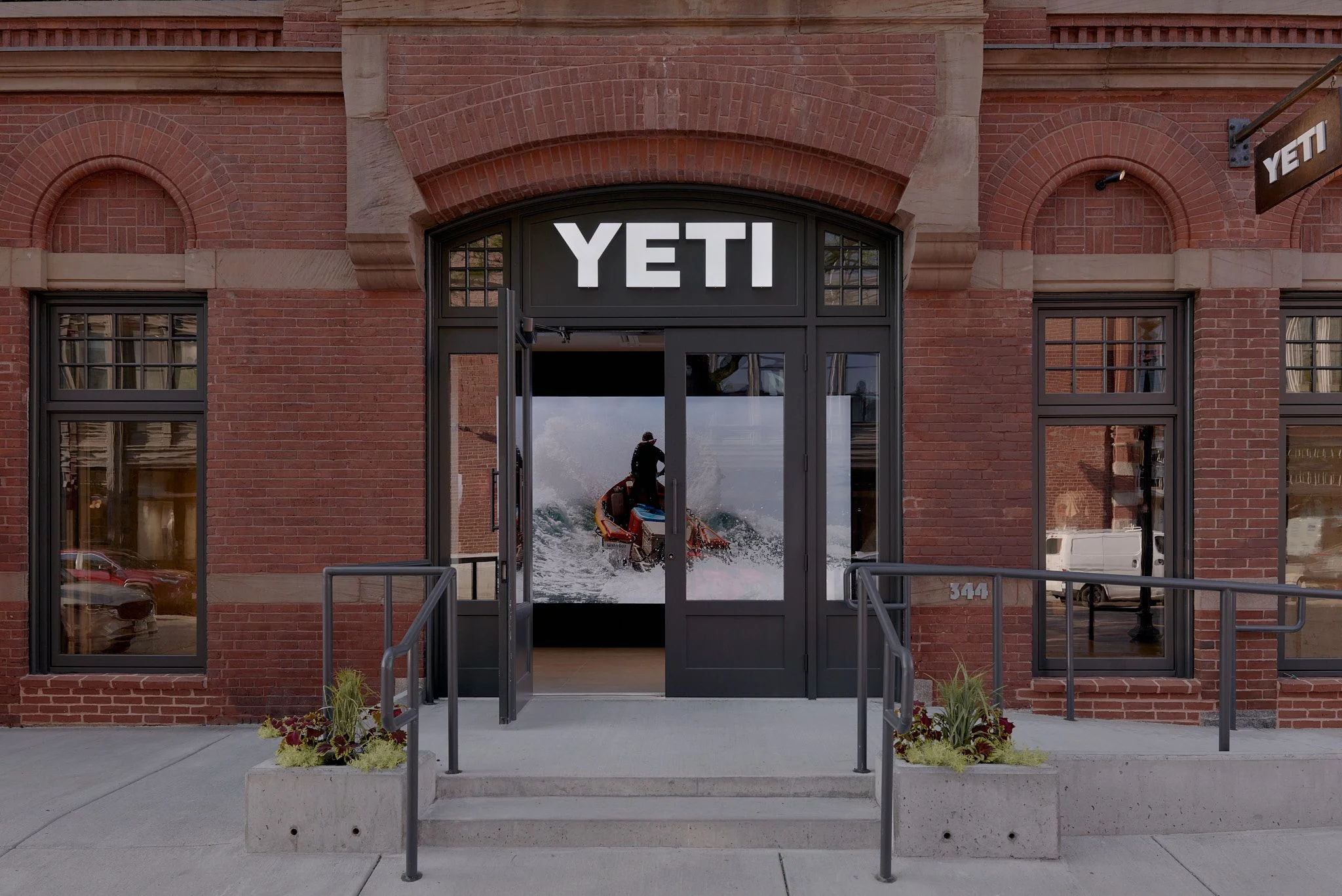 A modern YETI retail storefront features a dark-framed entrance with large glass doors set within a historic red brick building with arched windows and ornamental detailing; the storefront displays an action photograph of a person with outdoor gear against a snowy mountain landscape.