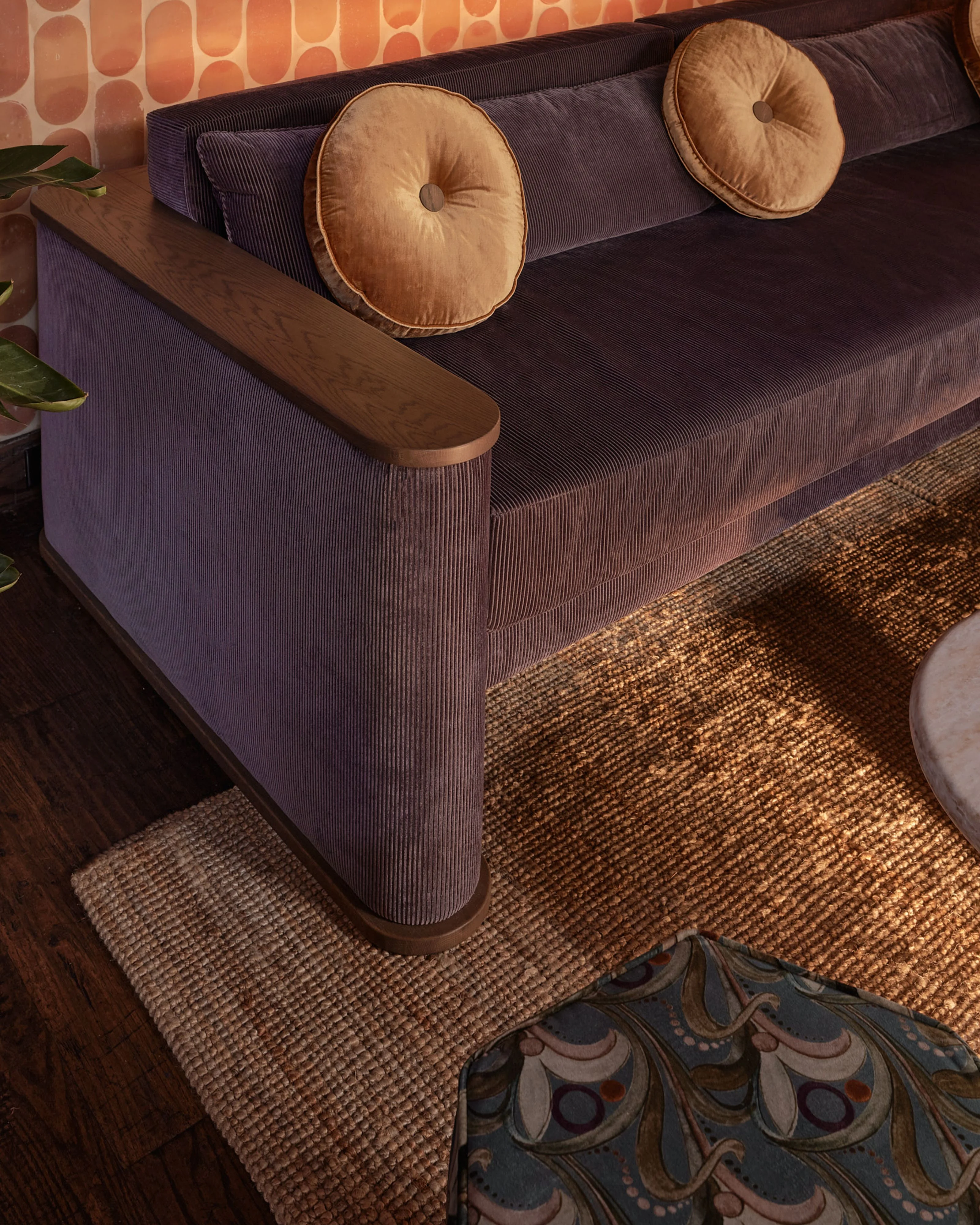 A modern living room features a dark purple ribbed sofa with round tan velvet bolster pillows and wooden armrests, positioned against a coral and cream geometric patterned wallpaper; warm natural light illuminates the space, which is anchored by a neutral textured area rug and dark hardwood flooring.