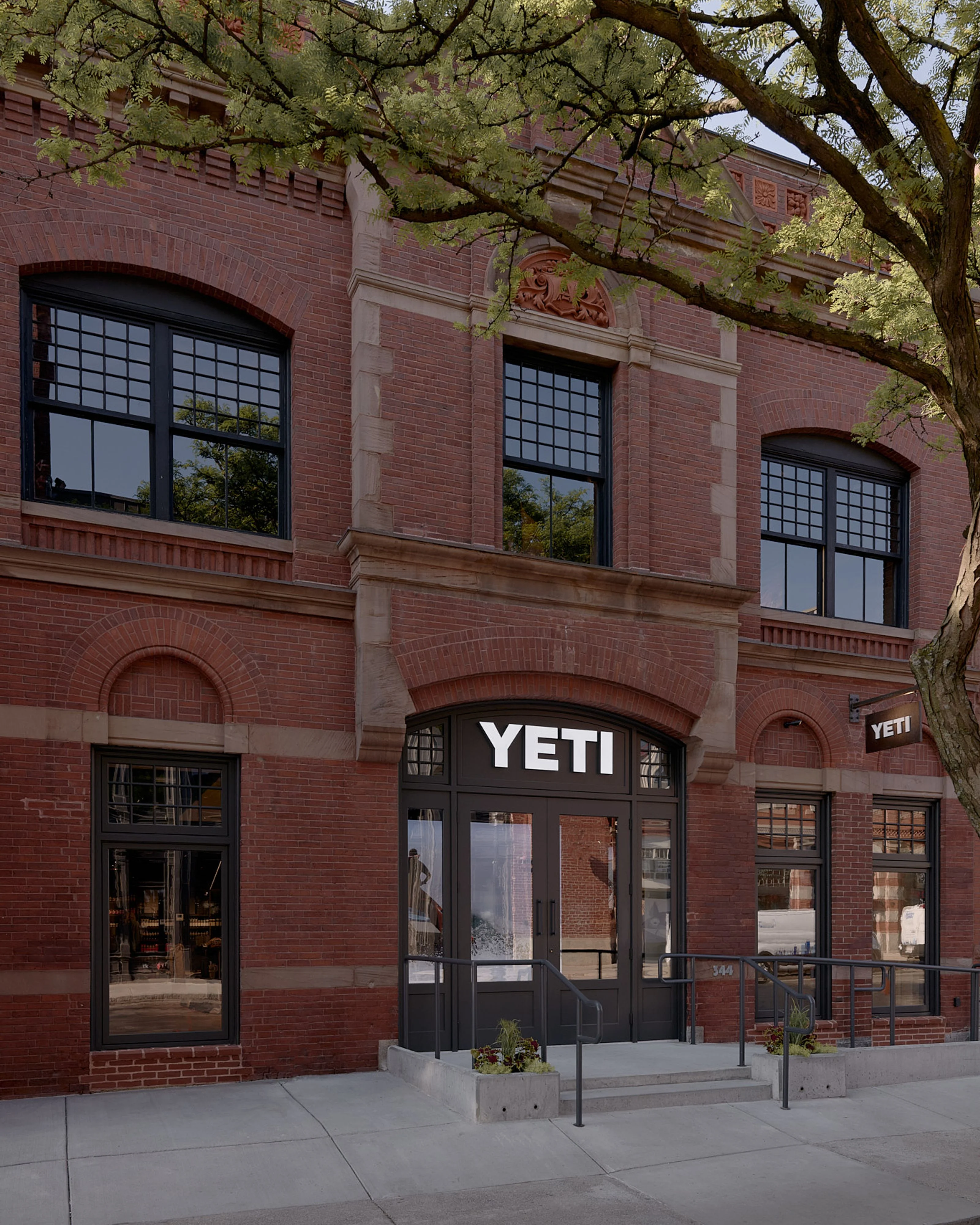 A modern YETI retail storefront occupies the ground floor of a historic red-brick building with arched windows and stone detailing, shaded by mature trees with delicate green foliage overhead. The storefront features dark-framed glass doors and windows with a minimalist design that contrasts with the building's classical Romanesque architecture.
