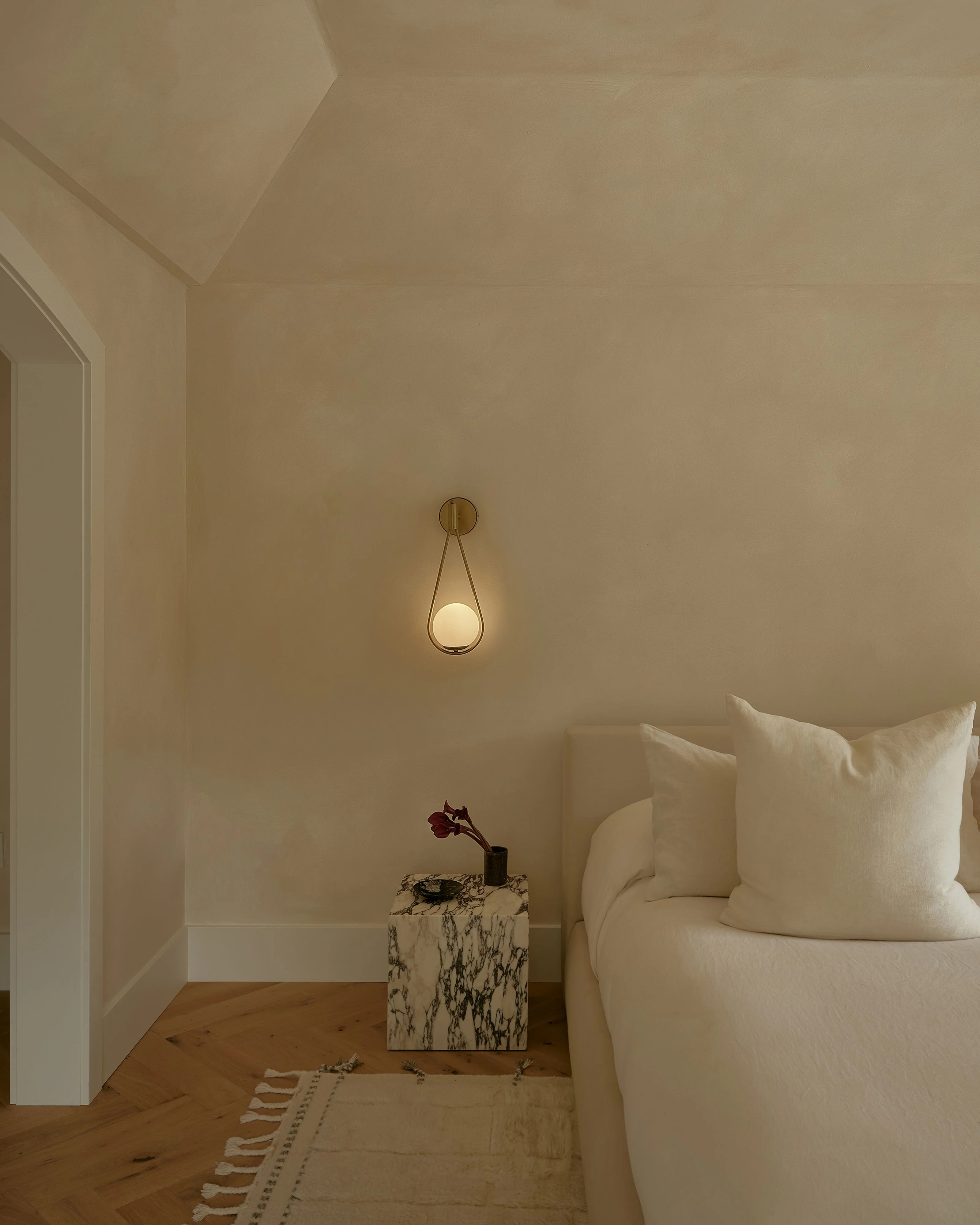 A minimalist bedroom with soft, neutral beige walls and wooden flooring features a cream-colored upholstered bed with plush pillows, a sculptural brass pendant light, and a marble-textured bedside table with a dark red flower stem. The serene, monochromatic space is completed by warm lighting and subtle architectural details including clean white molding and a patterned area rug.