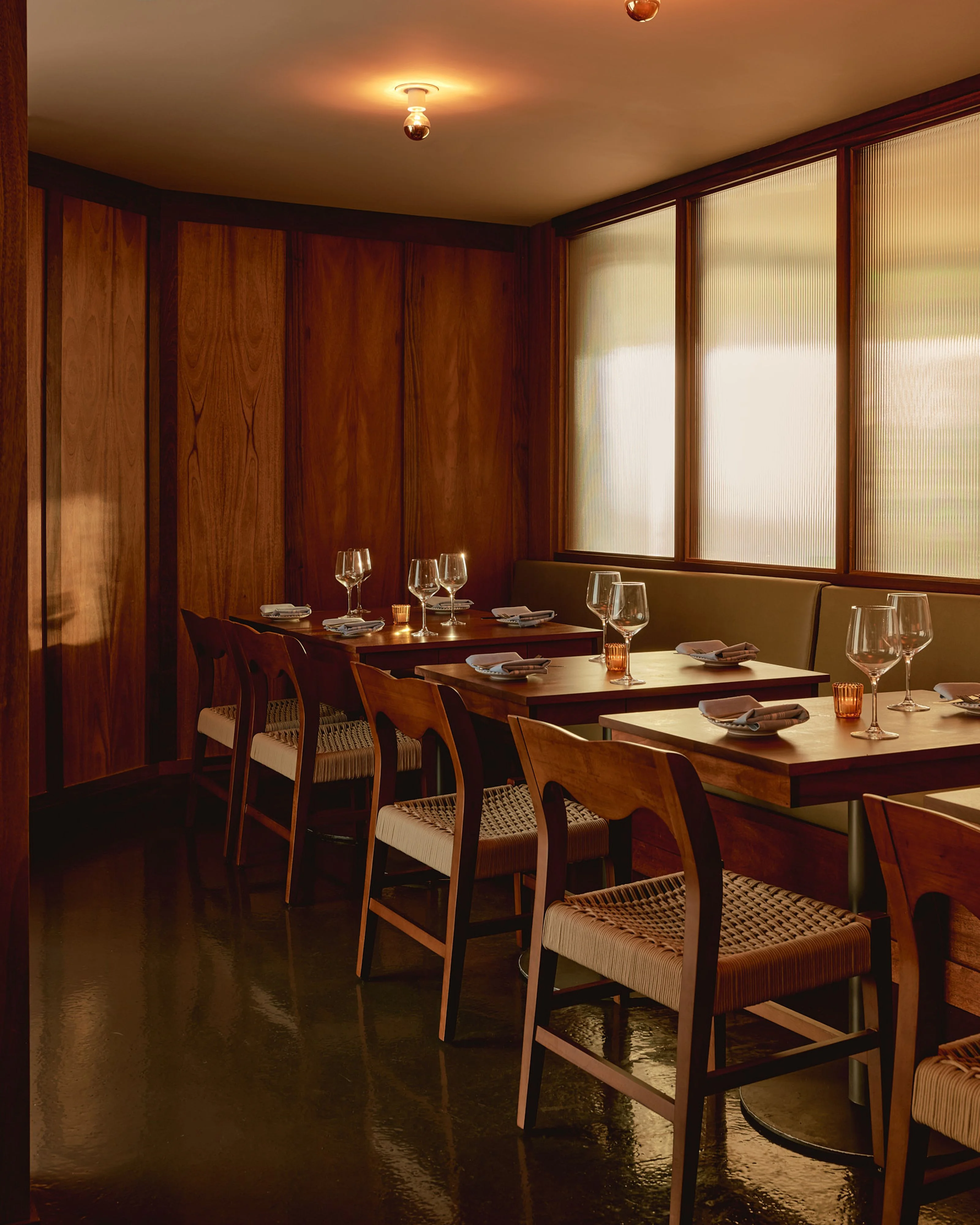 A warm, mid-century modern dining space features rich wood paneling and furniture with woven rush-seat chairs arranged at dark wood tables set with glassware and place settings; diffused natural light filters through large translucent windows along one wall, while warm overhead lighting creates an intimate, sophisticated ambiance.