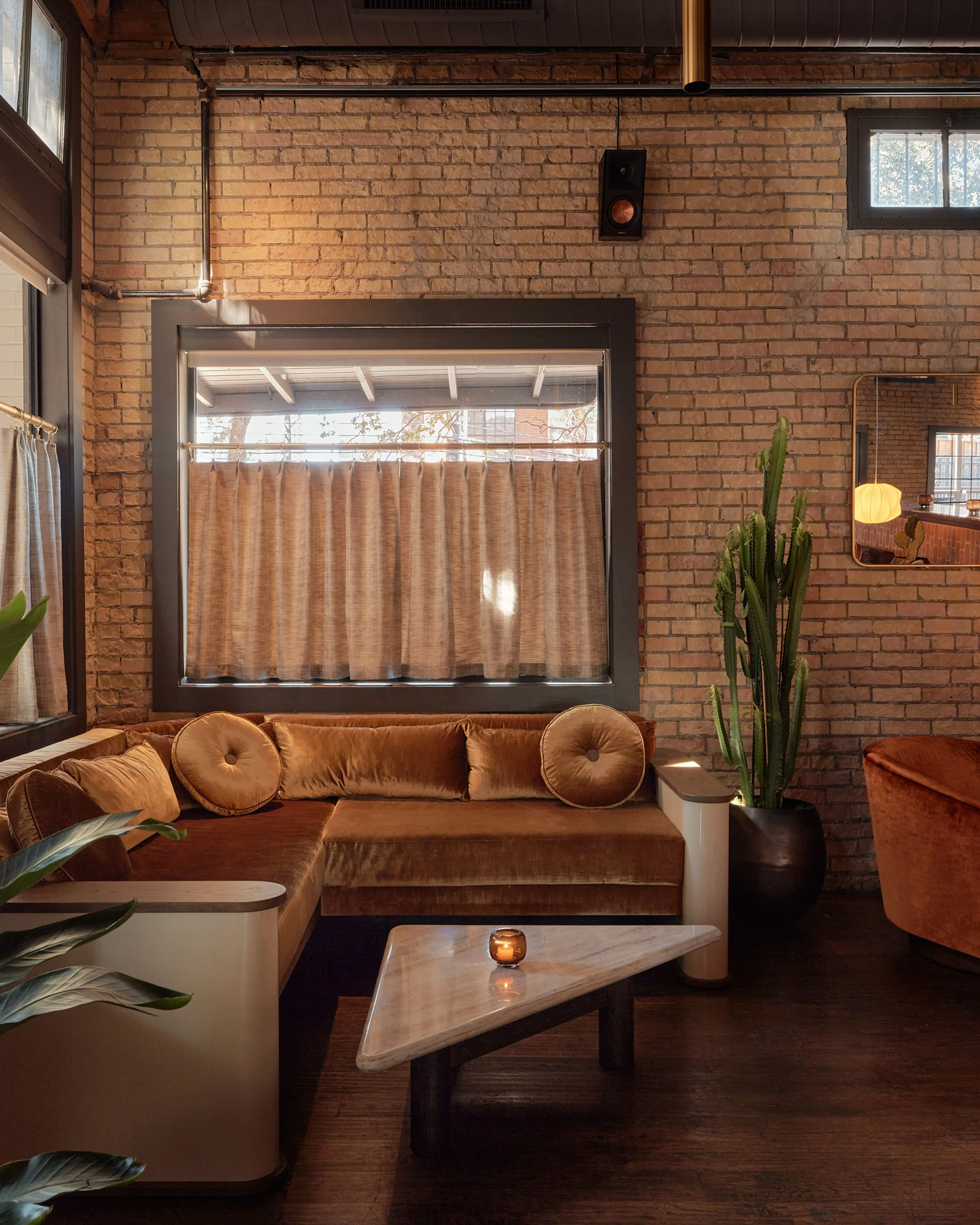 A warm, mid-century modern loft interior featuring a rust-colored velvet sectional sofa positioned beneath a large industrial window with tan linen curtains; exposed brick walls in rich terra-cotta tones are accented with dark wood beams, potted cacti, and minimalist furnishings.
