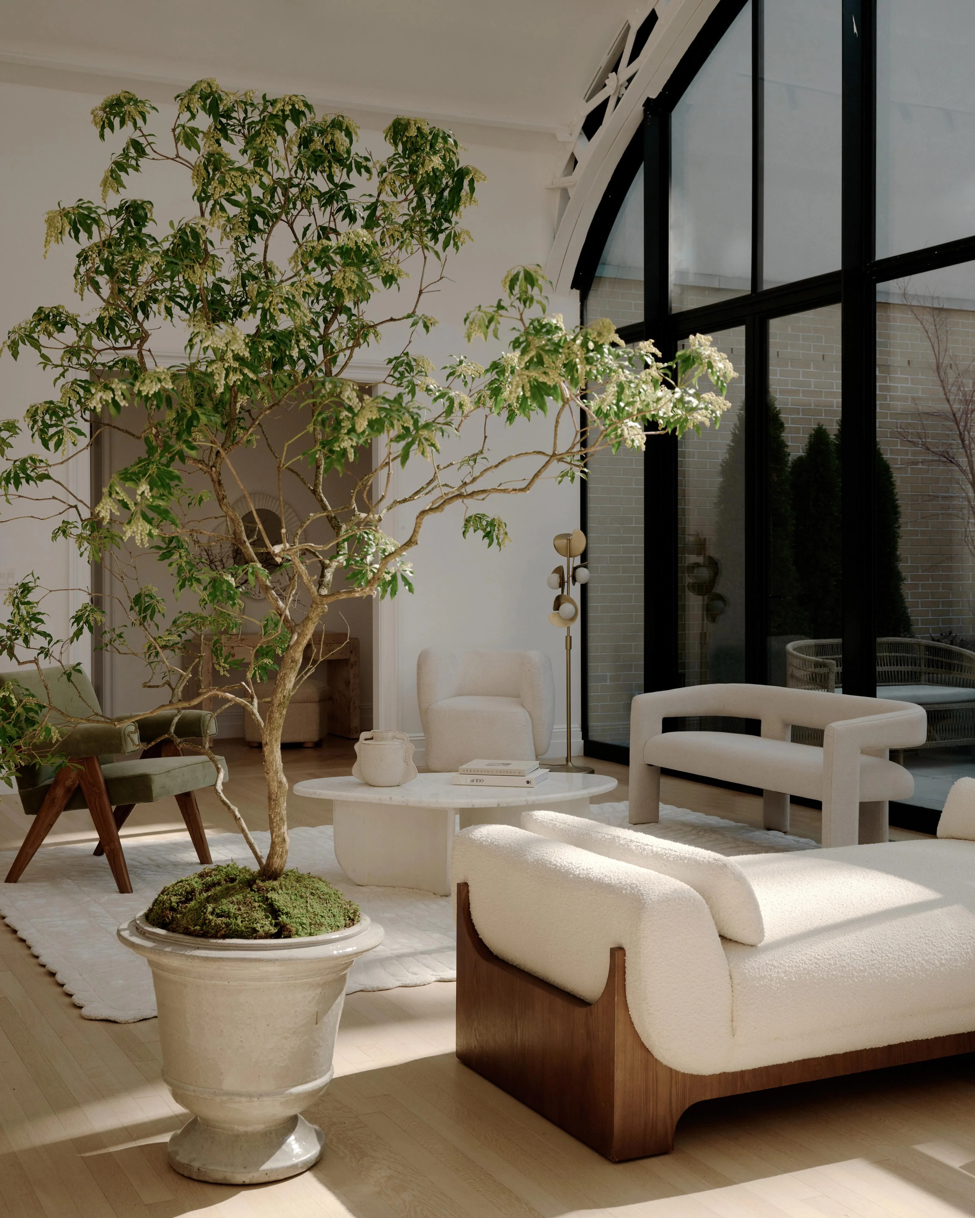A serene, minimalist interior living space flooded with natural light features cream-colored curved furniture with wood bases, potted greenery including a sculptural indoor tree and moss planter, and a black-framed glass wall system opening to a brick courtyard garden.