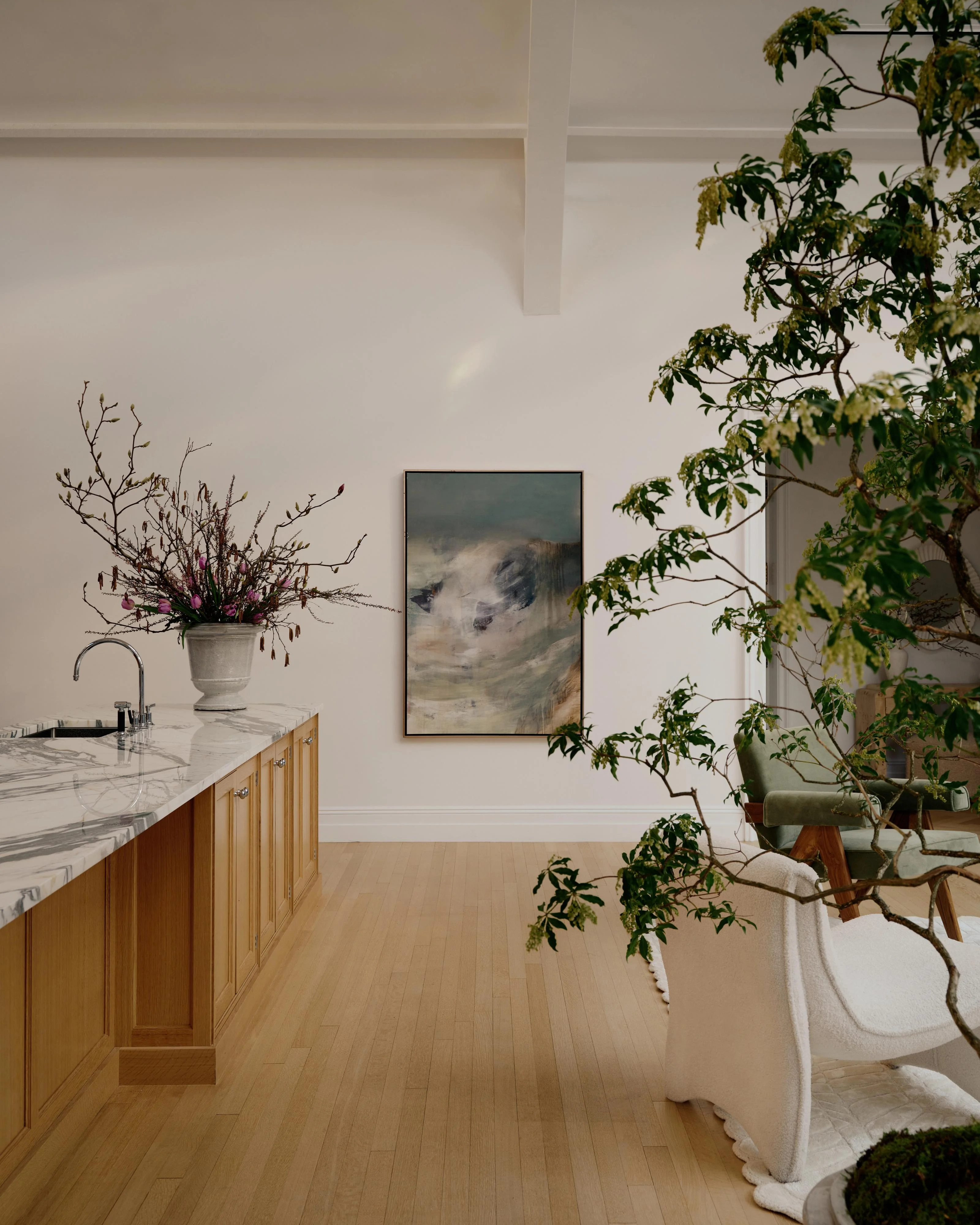 A minimalist kitchen and living space with white walls and light wood flooring features warm wooden cabinetry with a marble countertop on the left, while lush green plants and a dried floral arrangement in a cream pot frame the right side; a framed landscape artwork hangs on the white wall above a mid-century modern chair draped with a white blanket.