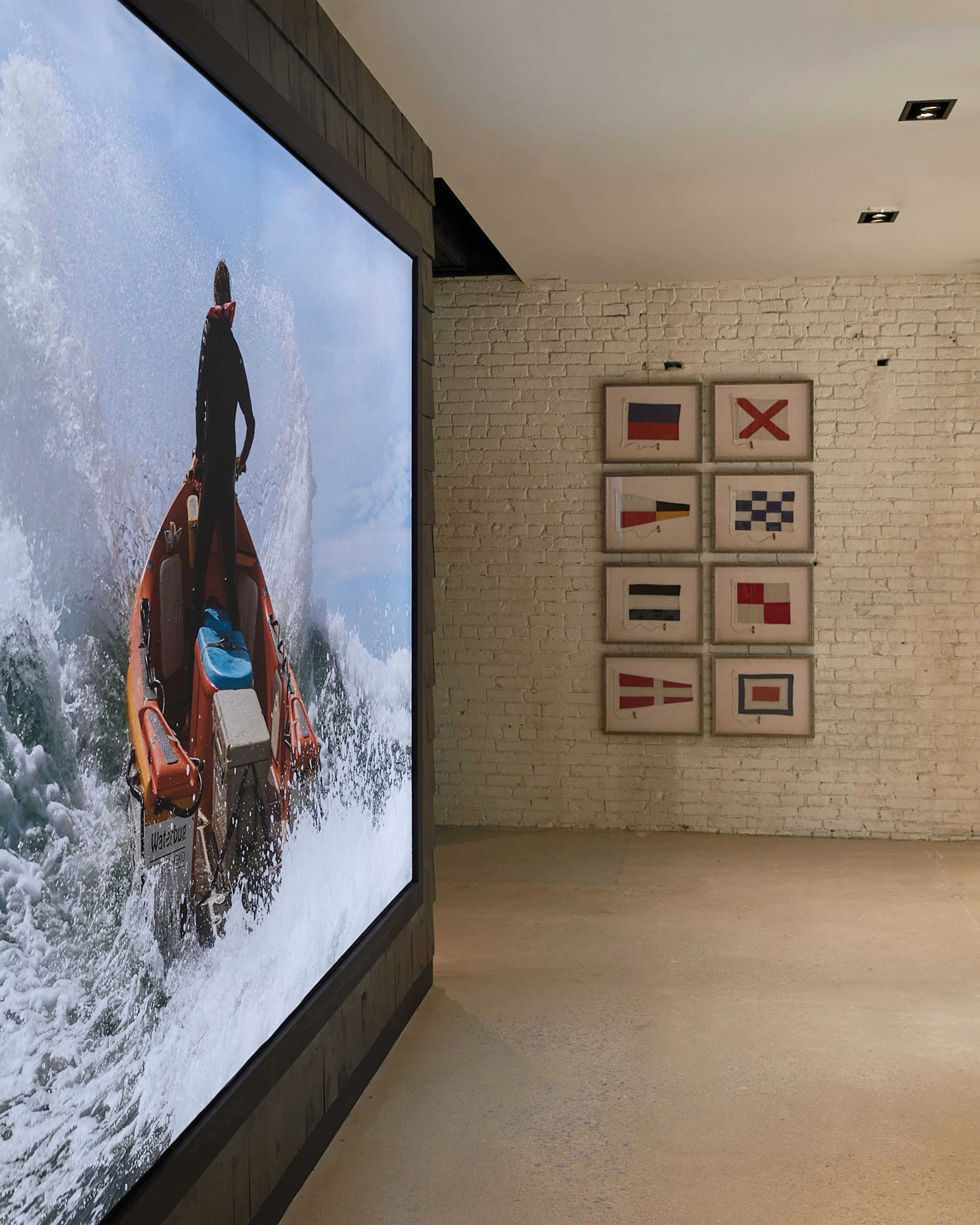 A minimalist interior space features a large digital display showing an orange lifeboat cutting through turbulent waves, while the opposite cream brick wall displays eight framed nautical flag symbols arranged in a grid, creating a maritime-themed gallery setting with polished concrete flooring and recessed ceiling lights.