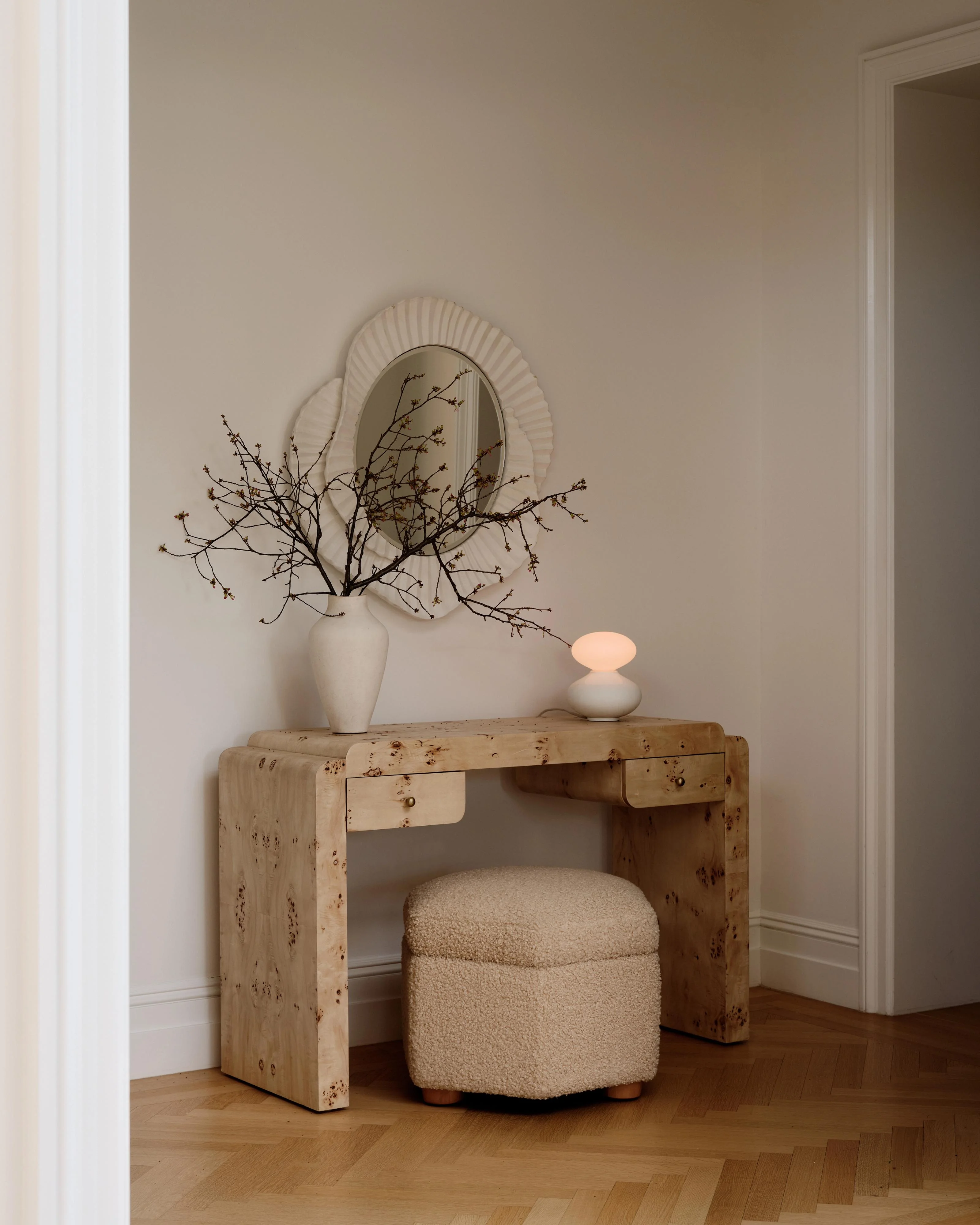A minimalist interior corner features a light wood desk with natural knots and grain patterns, topped with a white ceramic vase containing dried branches, a decorative mirror with a ribbed cream frame, and a small peach-toned sculptural lamp. A rounded beige upholstered ottoman sits beneath the desk on warm wood parquet flooring.