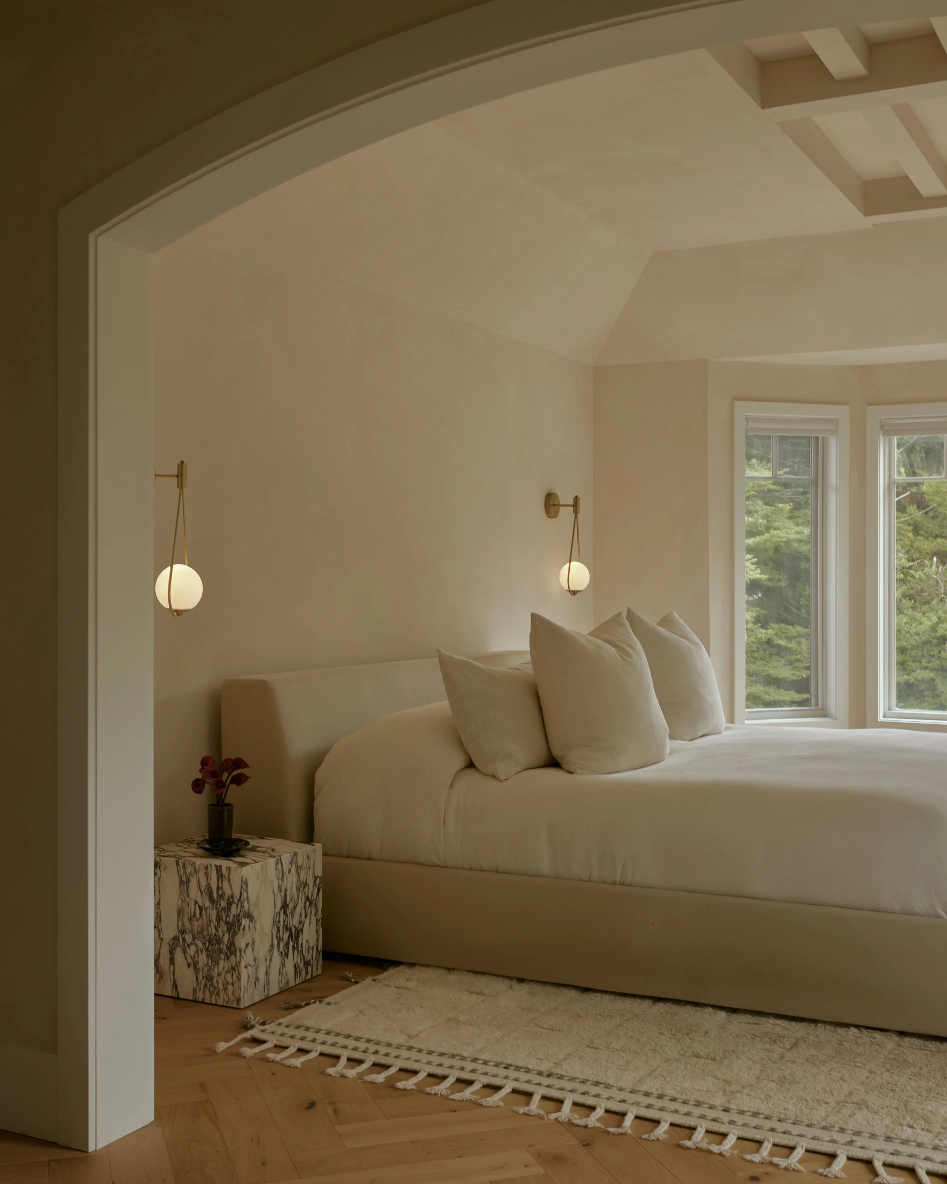 A serene, minimalist bedroom featuring a cream-upholstered bed with matching pillows, illuminated by two brass pendant lights with globe shades suspended above. The room is bathed in natural light from large windows overlooking greenery, with warm beige walls, wooden flooring, a marble side table with a burgundy flower arrangement, and a geometric woven area rug.