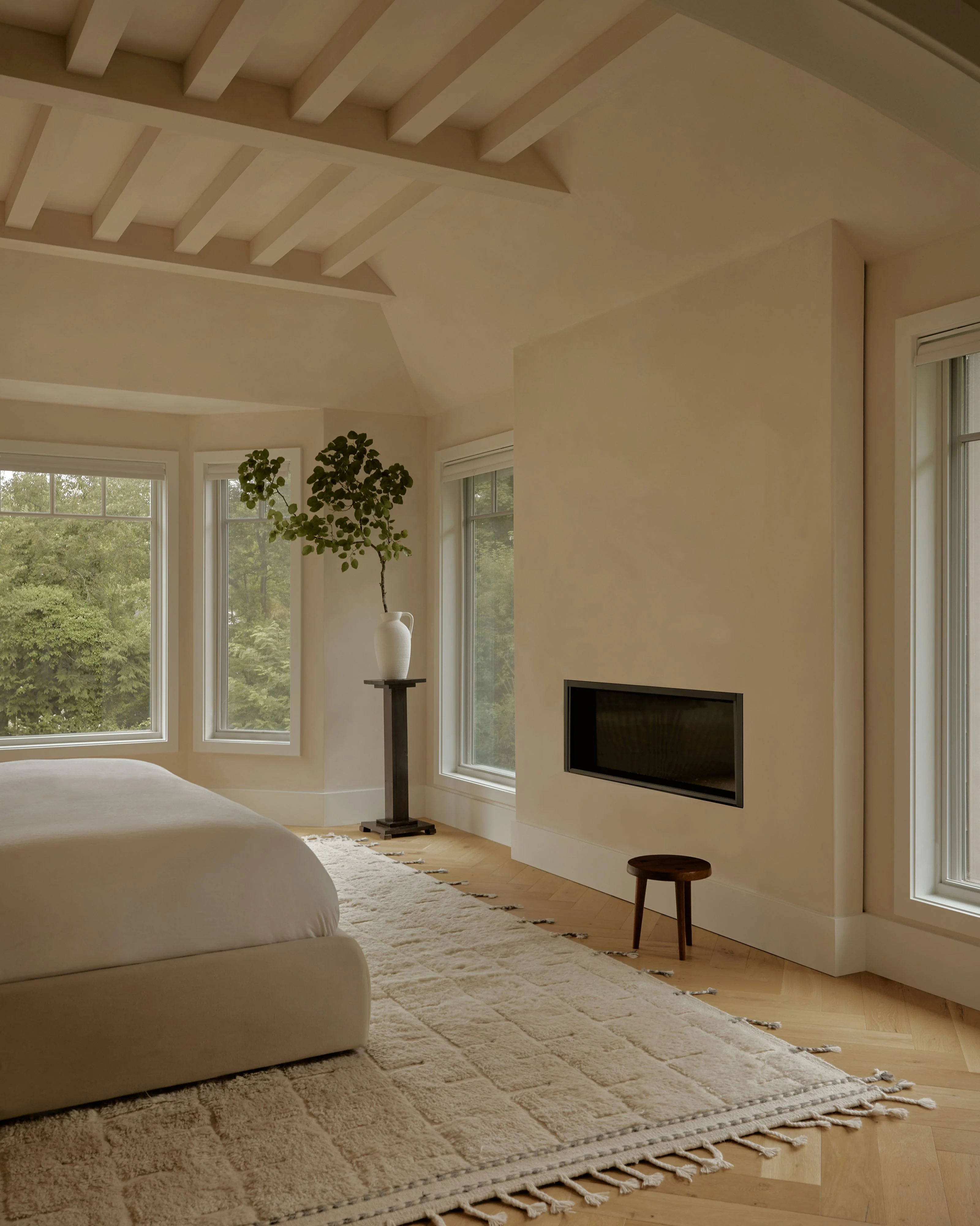 A minimalist, light-filled bedroom featuring cream-colored walls, exposed wooden ceiling beams, and light wood flooring, with a white curved sofa, beige woven area rug, wall-mounted flat-screen television, pedestal side table with greenery, and large multi-pane windows overlooking lush green foliage.