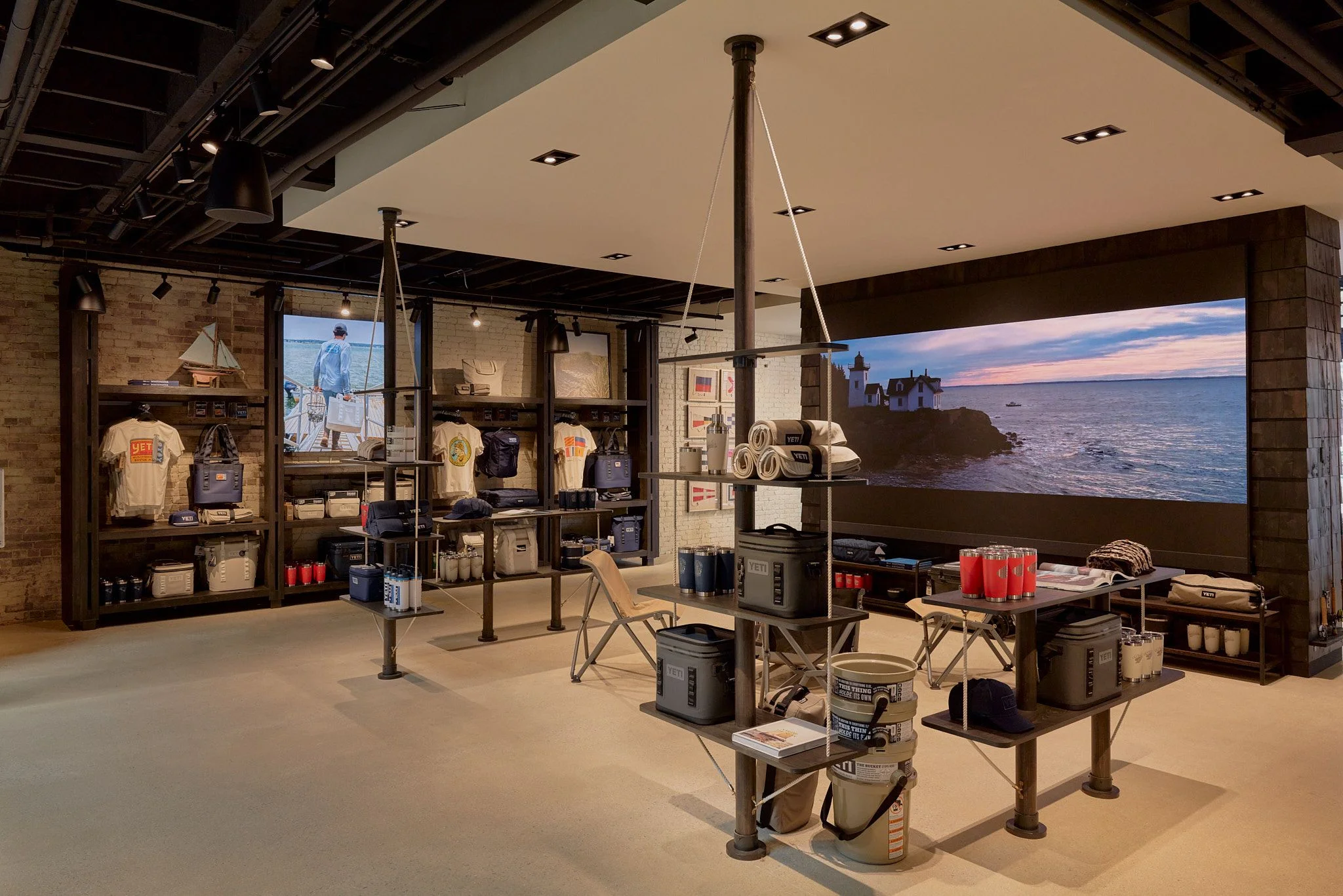 A modern retail showroom with industrial design features exposed brick walls, dark metal shelving units displaying apparel and gear, and tall metal support poles anchoring display tables throughout the open space; a large video screen on the right wall displays a coastal lighthouse scene at sunset.