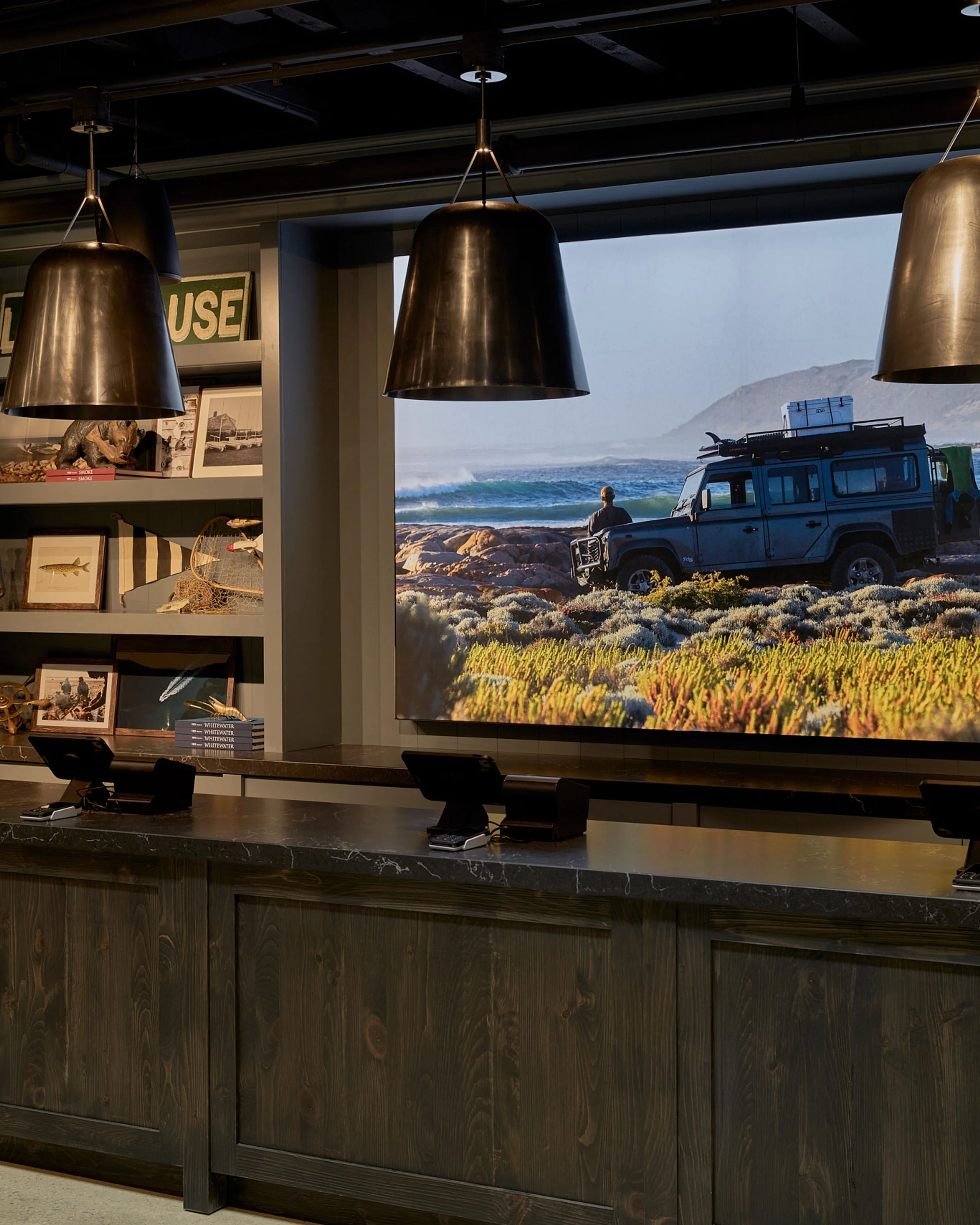 A modern interior space featuring a dark wood counter with three brass pendant lights suspended from a dark ceiling, facing a large display screen showing a scenic landscape with a blue adventure vehicle parked near coastal mountains. The left wall displays curated travel photography and memorabilia on shelves.