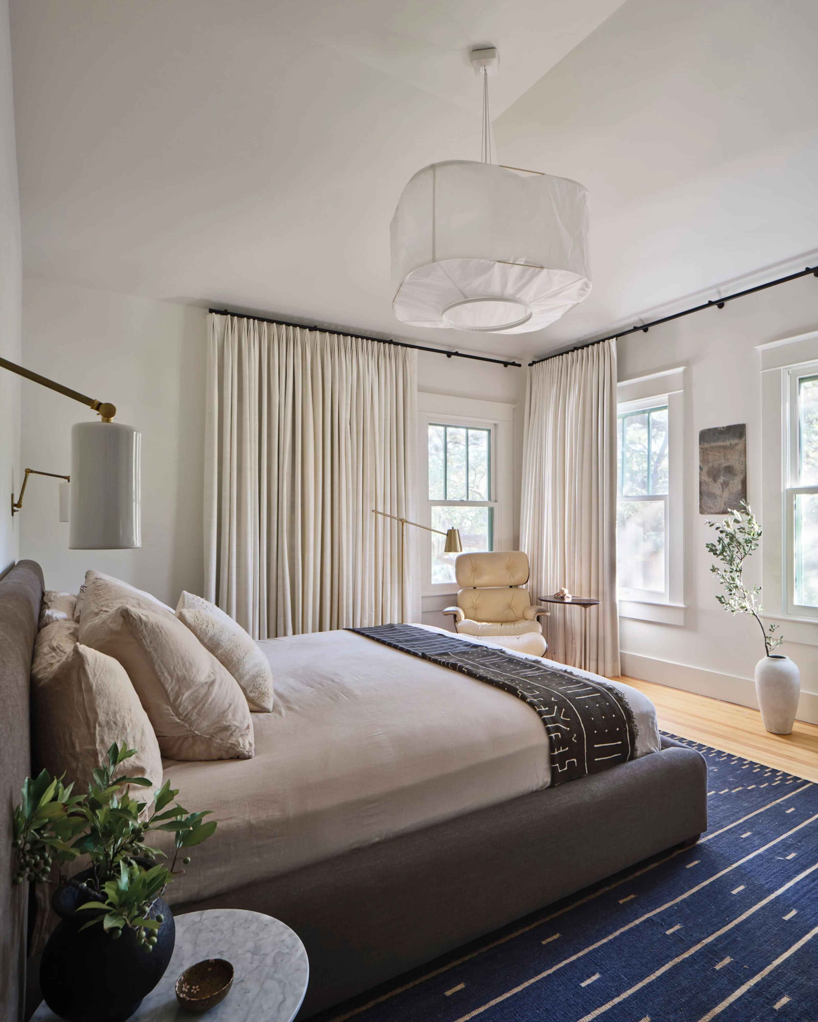 A serene, modern bedroom features crisp white walls and ceiling with a soft fabric pendant light centered overhead. The space showcases a gray upholstered bed dressed in neutral linens, flanked by cream floor-to-ceiling curtains, a camel-colored armchair, warm wood flooring, and a navy patterned area rug with gold geometric detailing.