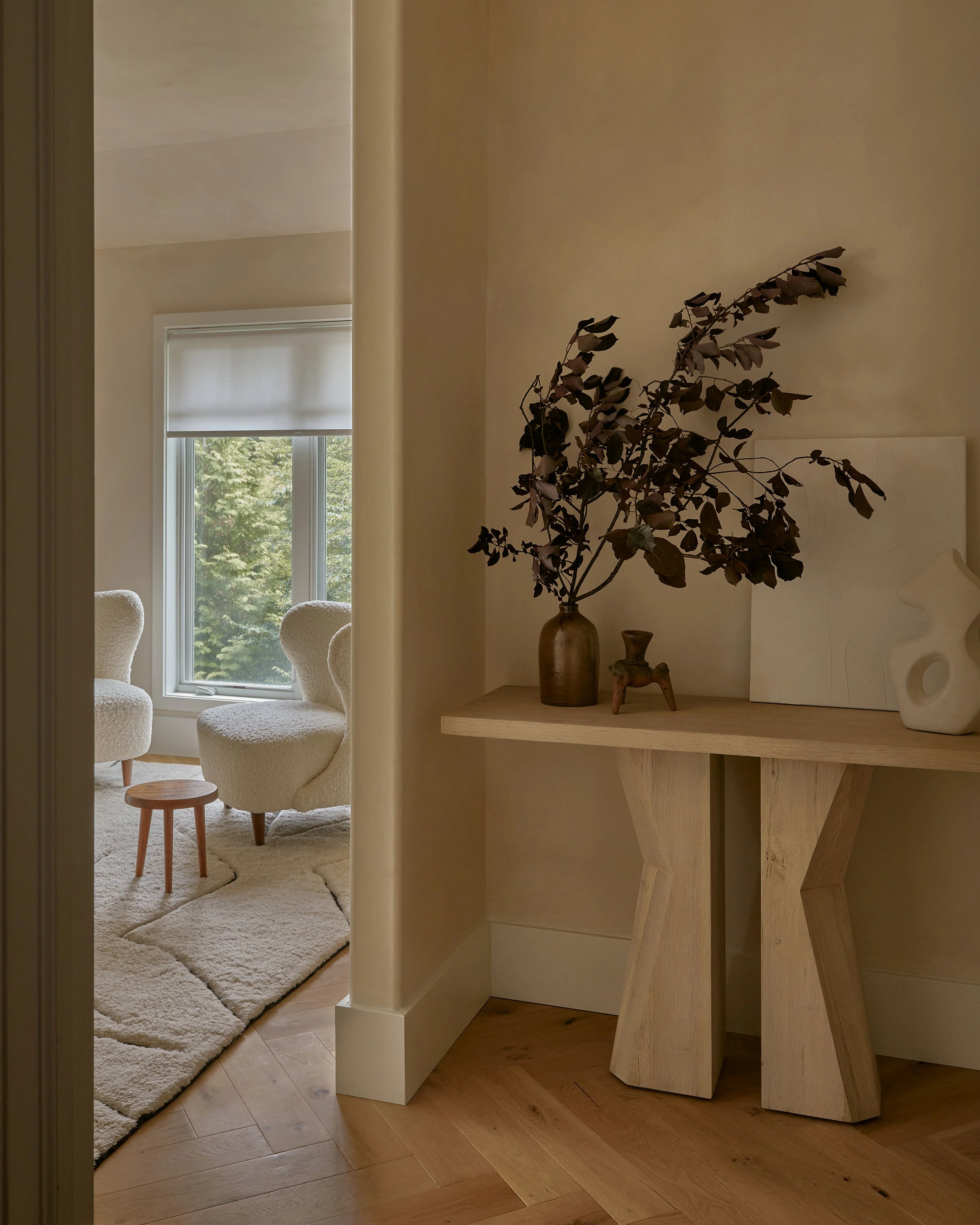 A serene, minimalist interior features a light wood console table adorned with dried botanicals in amber glass and ceramic vessels, positioned in a warm beige room with clean white walls and natural light. Through an open doorway, cream-colored sculptural chairs and a small wooden side table are visible near a large window overlooking greenery.