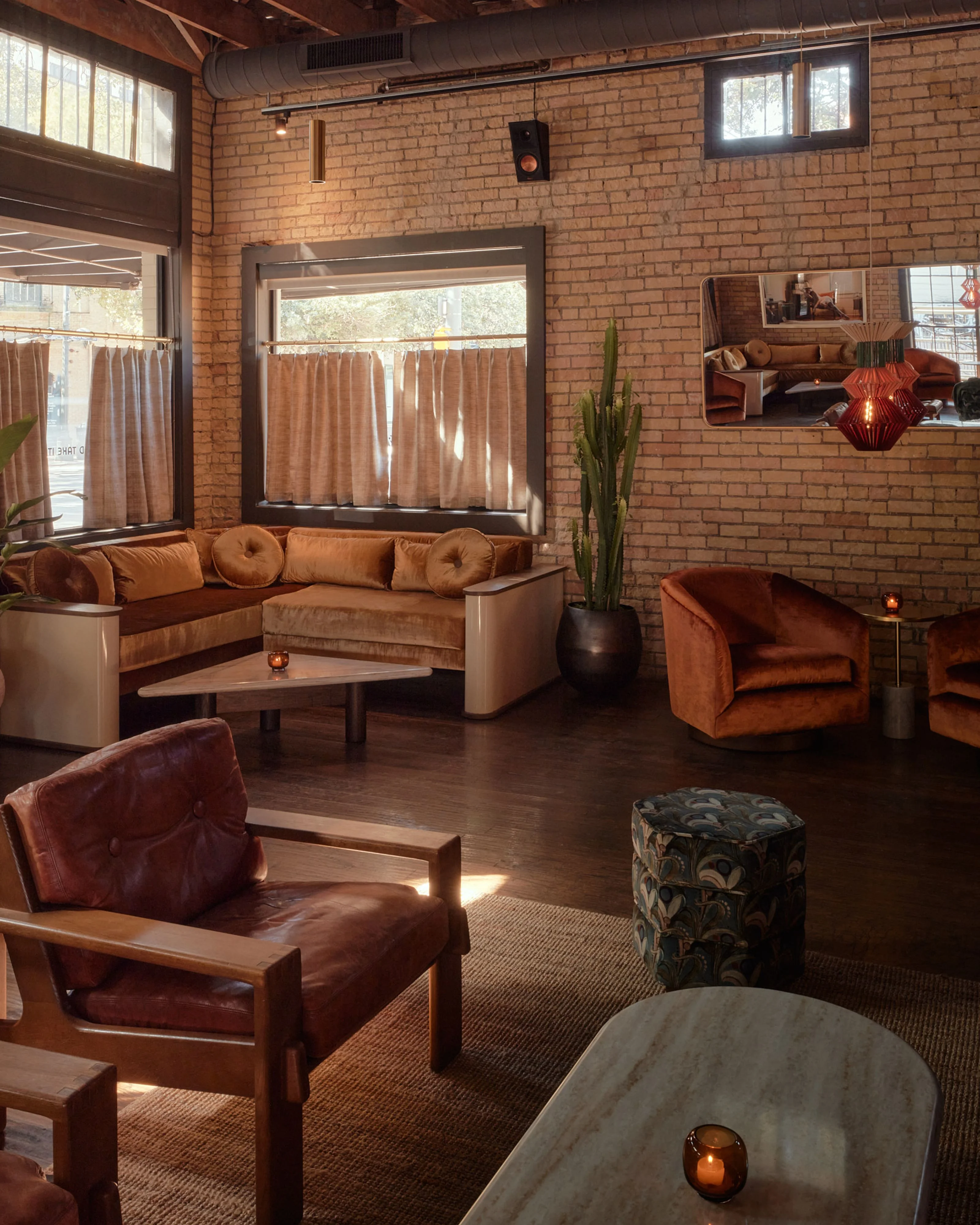 A warm, mid-century modern lounge space with exposed red brick walls, featuring a curated arrangement of cognac and rust-toned velvet and leather furniture, natural wood accents, and large industrial windows with neutral curtains that flood the dark wood flooring with natural light.