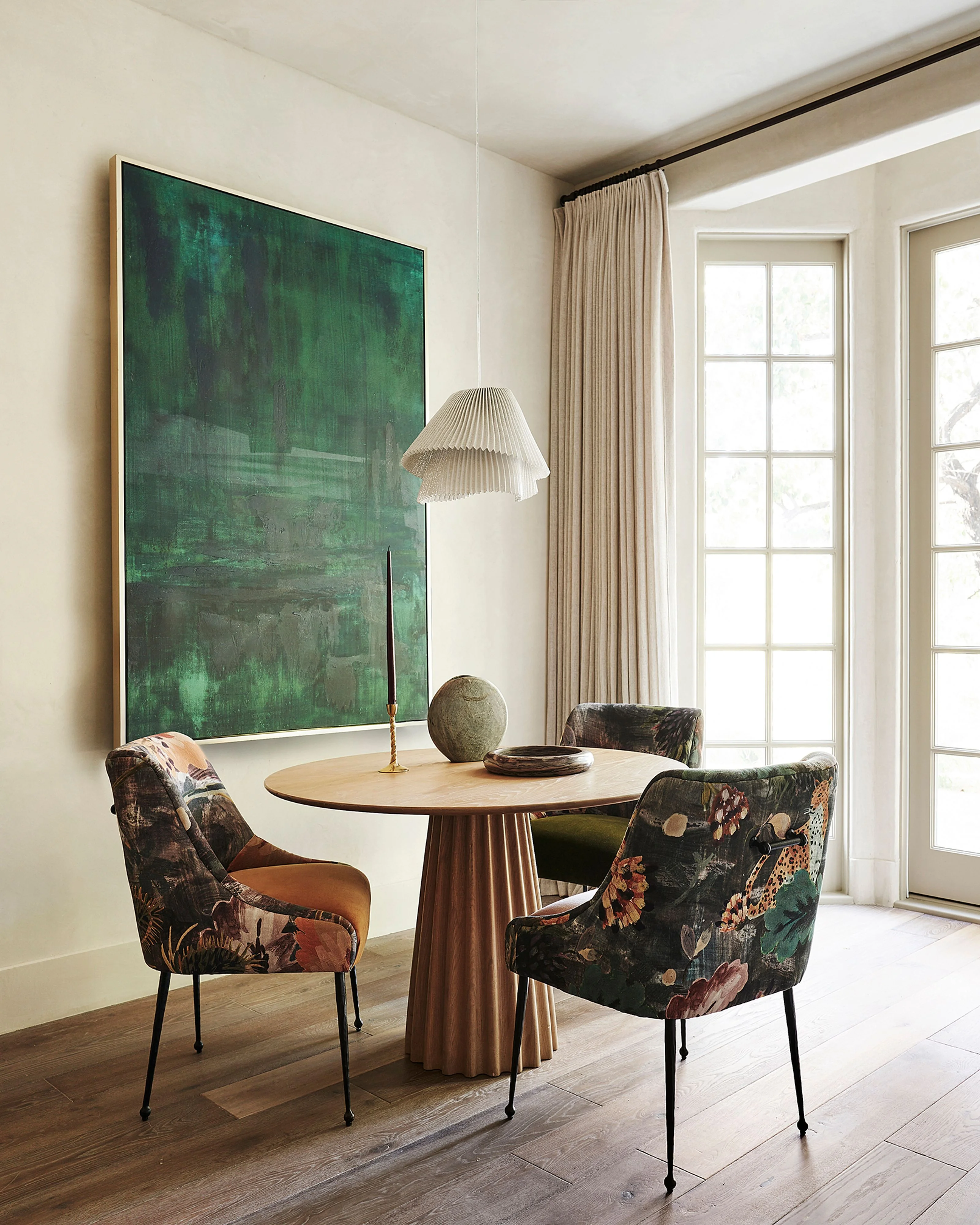 A bright, minimalist interior space features a round wooden table with fluted pedestal base surrounded by two floral-patterned upholstered chairs with metal legs, positioned beneath a large abstract green artwork with brass frame. Natural light floods through tall multi-pane glass doors and windows dressed with flowing cream curtains.