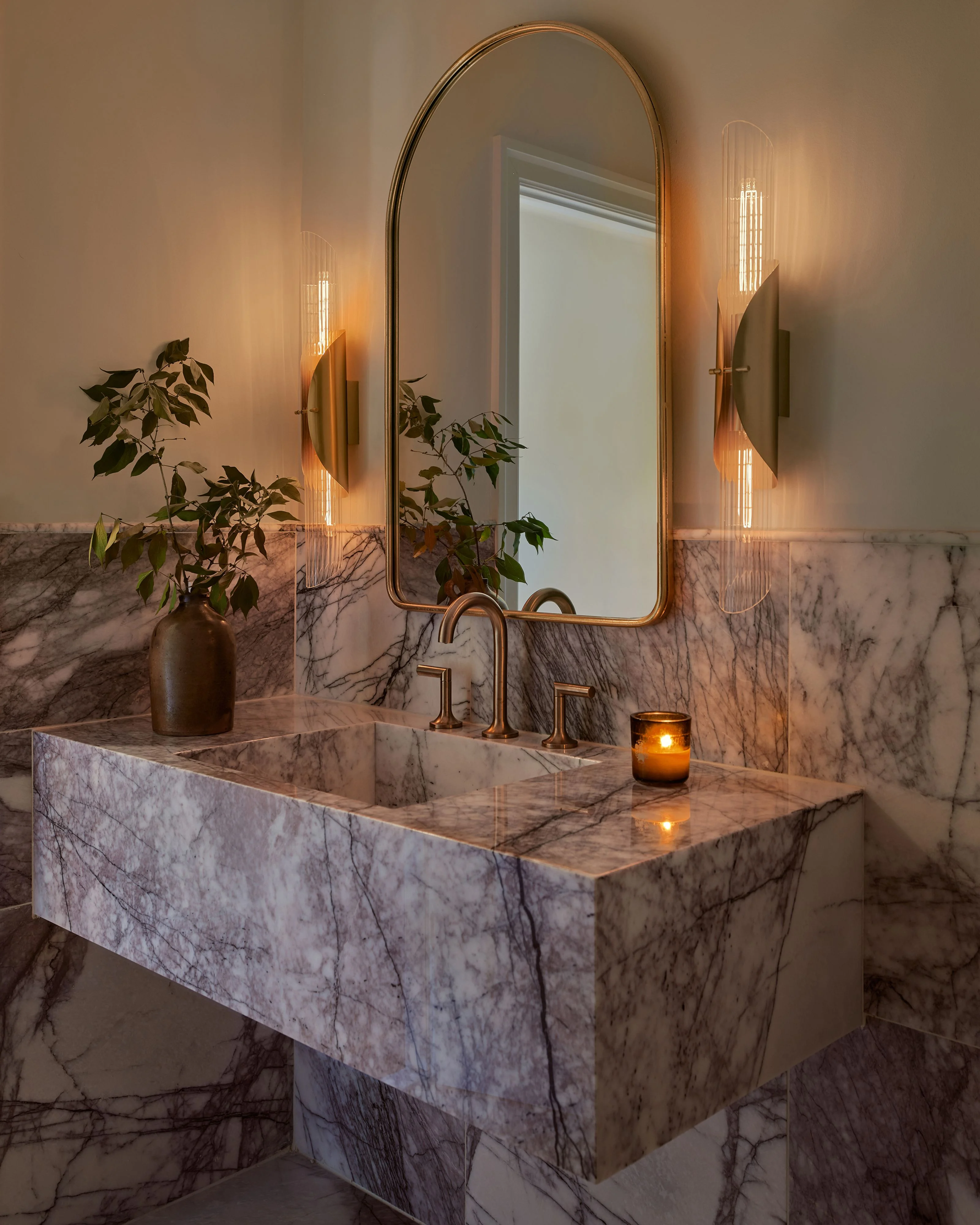 A luxurious bathroom vanity features a sculptural marble sink and countertop with dramatic veining, topped with warm brass fixtures and an arched brass-framed mirror. The serene, spa-like space is illuminated by elegant vertical linear wall sconces that cast a warm glow across taupe walls, complemented by potted greenery and flickering amber candles.