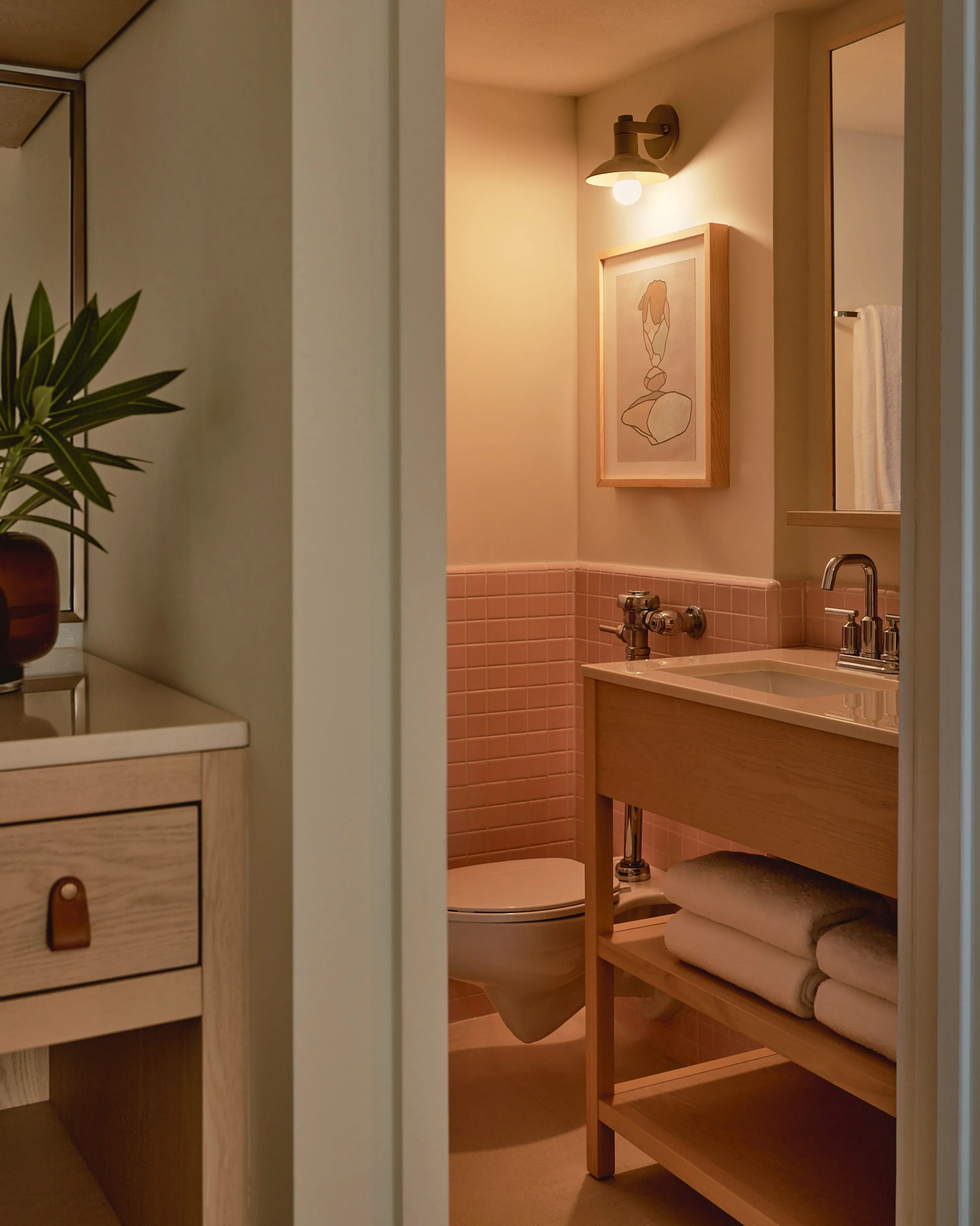 A warm, modern bathroom with peachy-coral tones features a vanity with a vessel sink, subway tile wainscoting, a wall-mounted brass sconce, and framed line art above the toilet; rolled towels and potted greenery add natural accents to the minimalist wood cabinetry and soft ambient lighting.