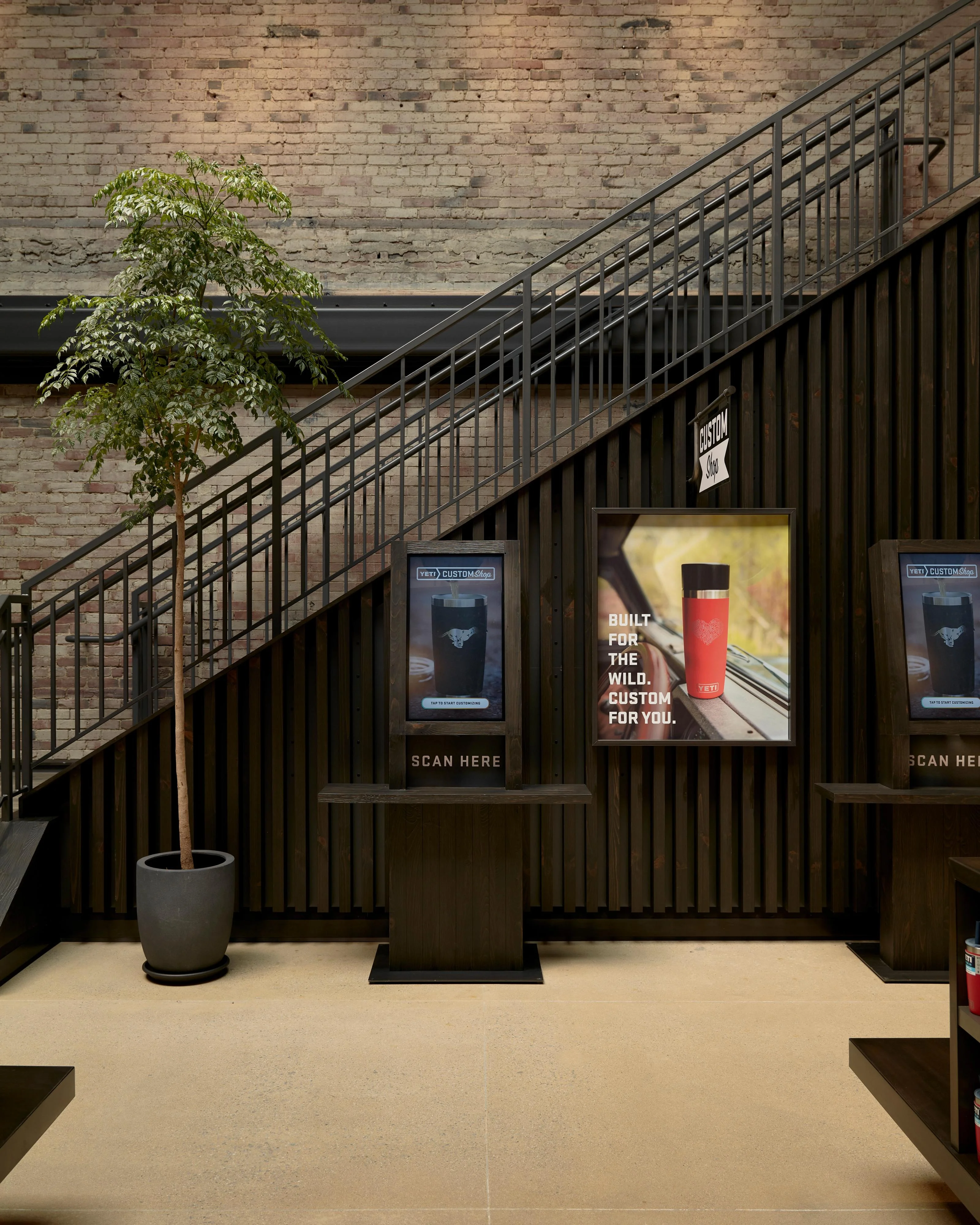 A modern industrial retail space featuring a diagonal black metal staircase with geometric railings against an exposed brick wall, with a light concrete floor, potted green plant, and illuminated product displays advertising customizable outdoor gear mounted on dark corrugated metal panels.