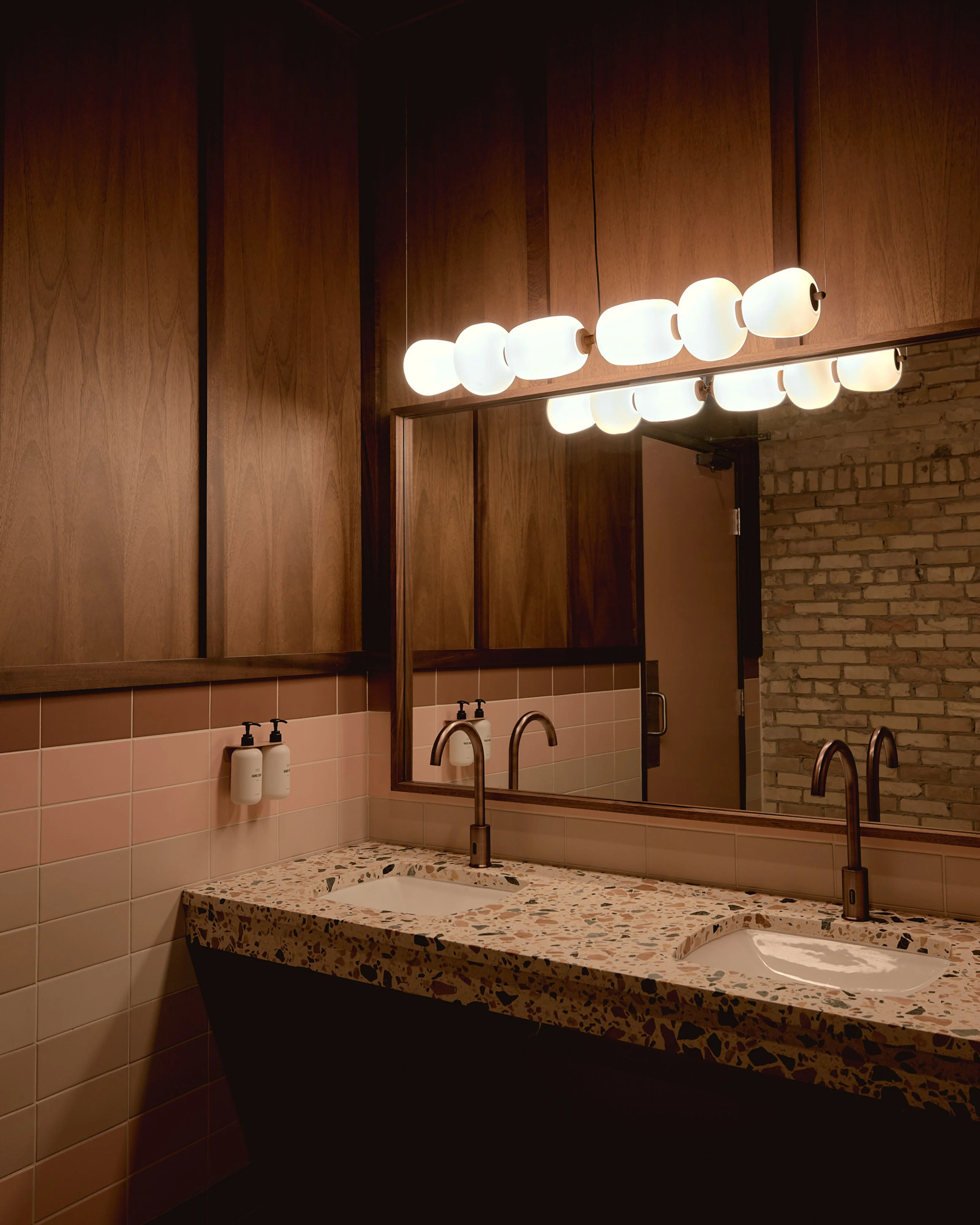 A warm, elegantly lit bathroom features rich wood paneling above a blush-pink tile backsplash, with a long terrazzo vanity countertop and bronze fixtures illuminated by a striking mid-century modern globe pendant light fixture. Exposed brick walls on the right and a large framed mirror complete the sophisticated, warm-toned design.