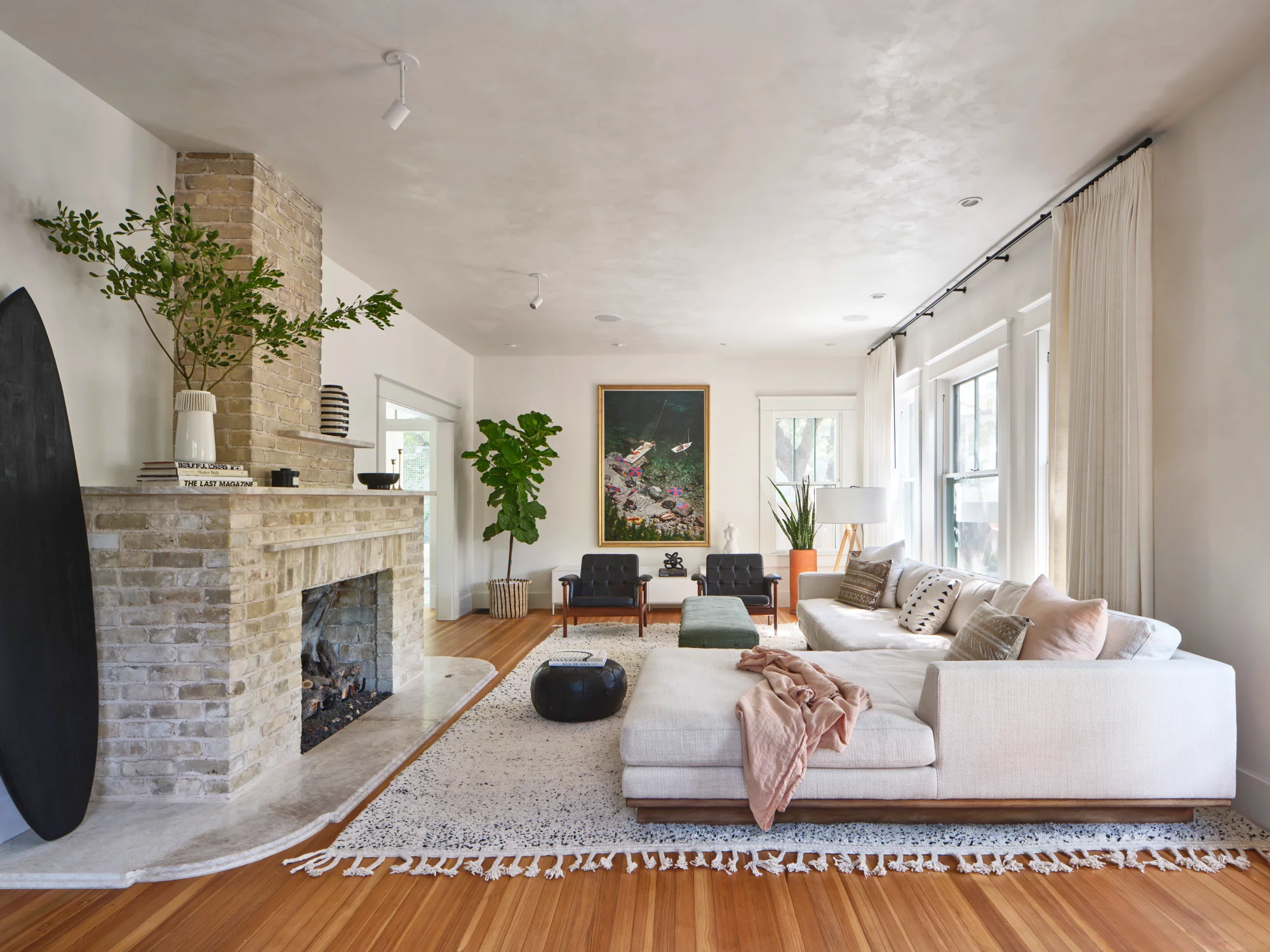 A bright, modern living room features a cream-colored sectional sofa anchoring a neutral color palette, with warm wood flooring, a textured area rug, and a prominent tan brick fireplace; tall windows with cream curtains flood the space with natural light, while mid-century modern seating, potted greenery, and a framed botanical artwork create a serene aesthetic.