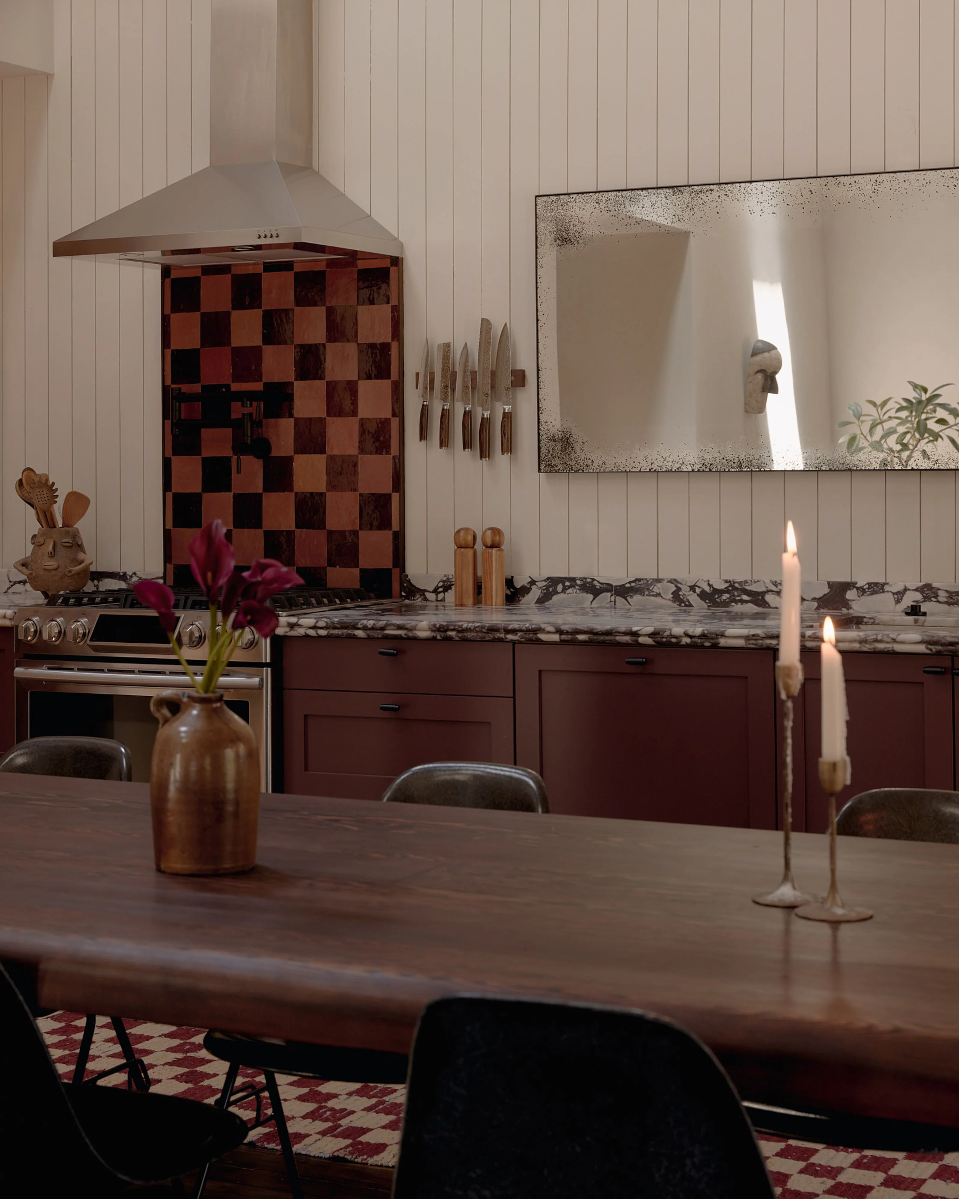 A modern kitchen and dining space with vertical shiplap walls features a dark wood dining table, burgundy cabinetry with a marble countertop, and a dramatic checkerboard-patterned accent wall behind a range hood, accented by warm candlelight and curated decor including a speckled mirror, wooden vessels, and sculptural pieces.