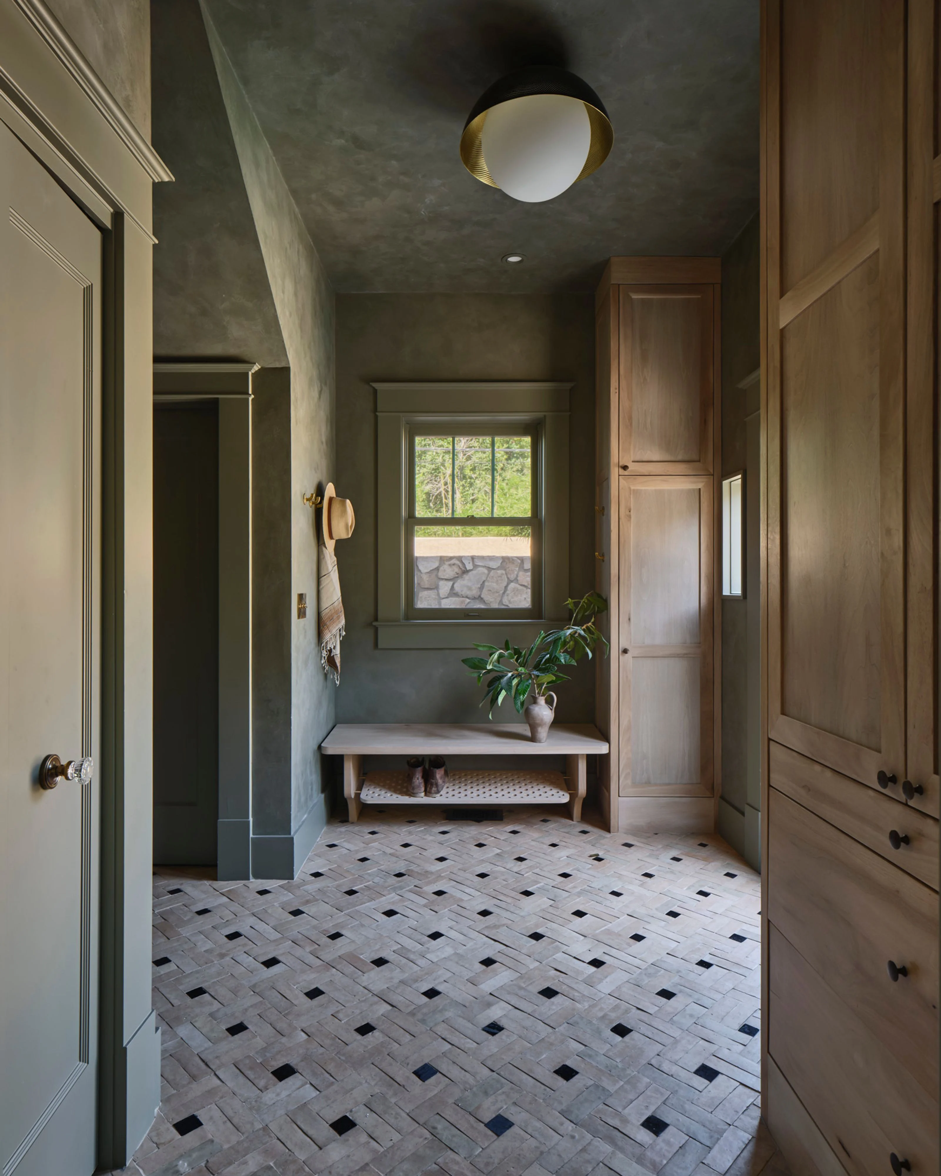 A sophisticated bathroom hallway features sage green walls with a polished concrete ceiling, herringbone tile flooring with black diamond accents, and warm wood cabinetry. Natural light streams through a large window above a minimalist console table with a potted plant, while a brass and black ceiling fixture completes the elegant, spa-like aesthetic.
