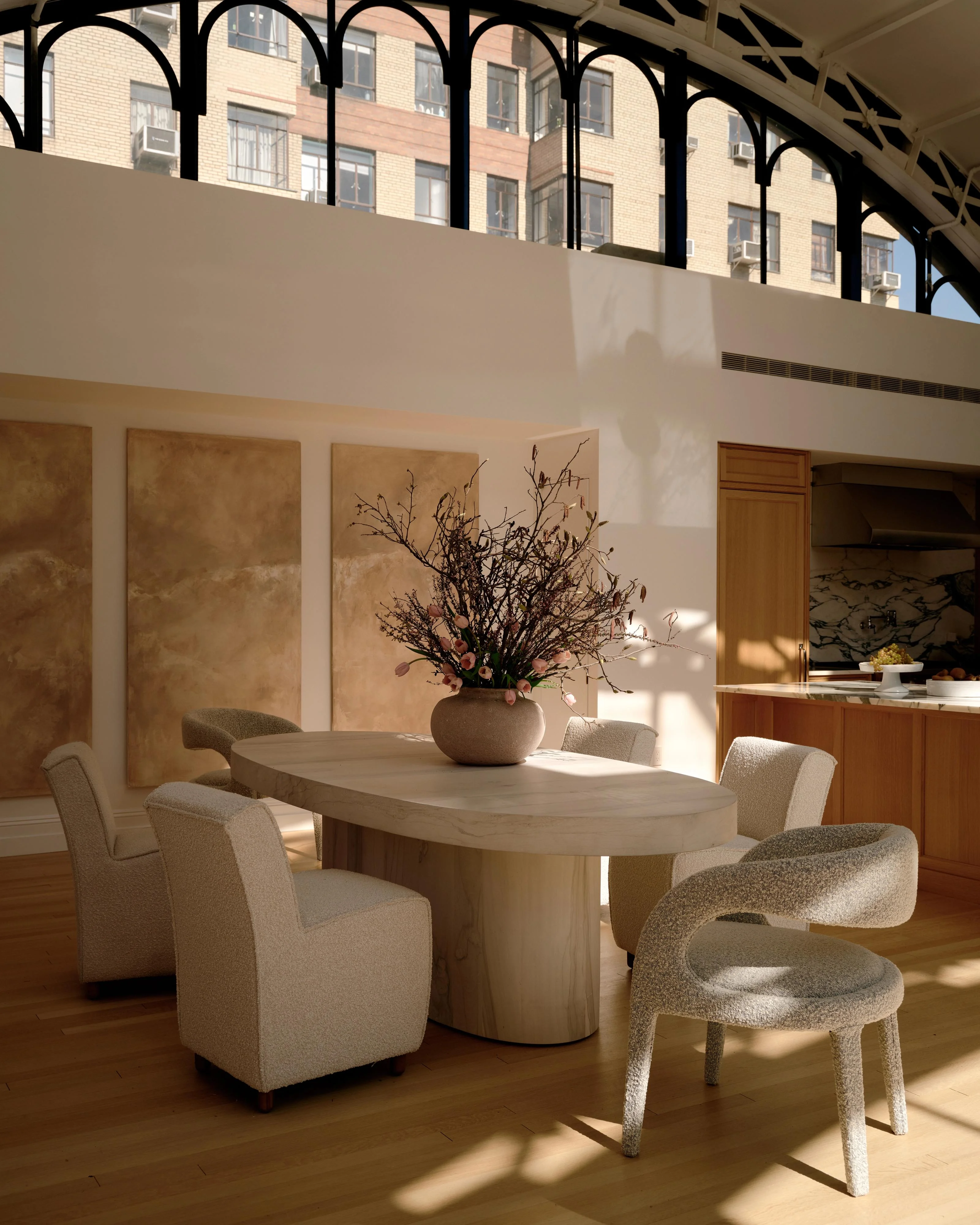 A bright, minimalist dining space featuring a round light wood table surrounded by curved cream-colored upholstered chairs, with a spherical ceramic vase of dried branches as a centerpiece, bathed in warm sunlight from large arched windows; the room showcases clean lines, neutral tones, warm wood cabinetry, and geometric wall panels against exposed brick urban views.