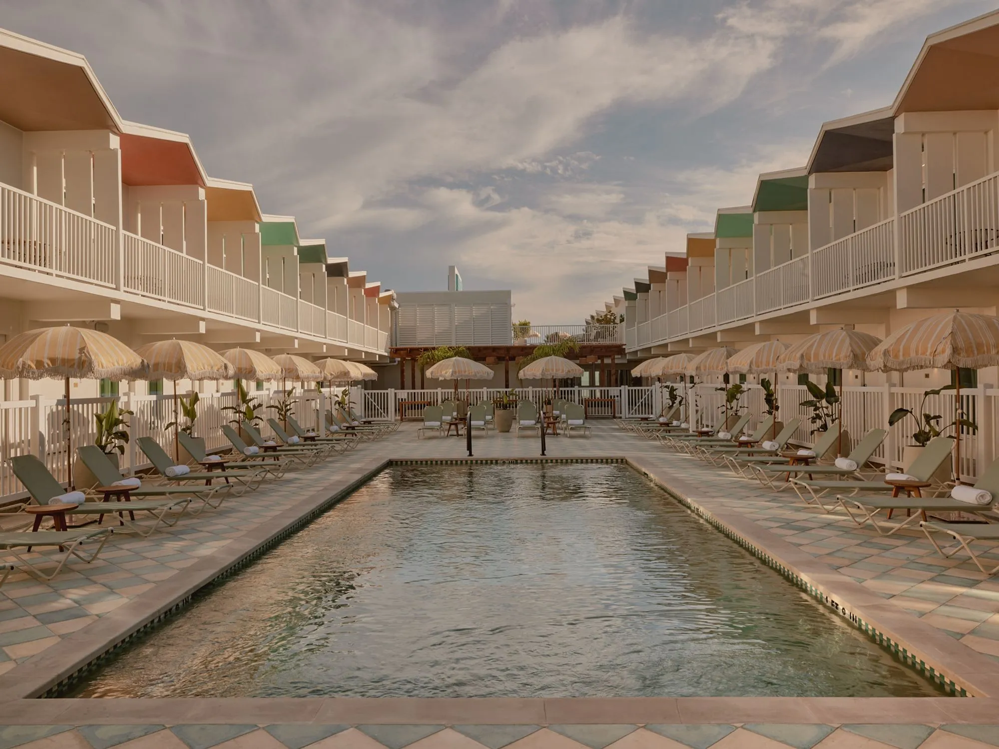 A symmetrical beachfront resort courtyard features a rectangular swimming pool flanked by rows of striped umbrellas and lounge chairs, with cream-colored beach houses featuring colorful roofs lining both sides and white railings under a soft, cloudy sky. The serene, nostalgic mid-century aesthetic is enhanced by manicured landscaping, terracotta paving, and a cohesive design palette of warm neutrals and vintage-inspired beach architecture.