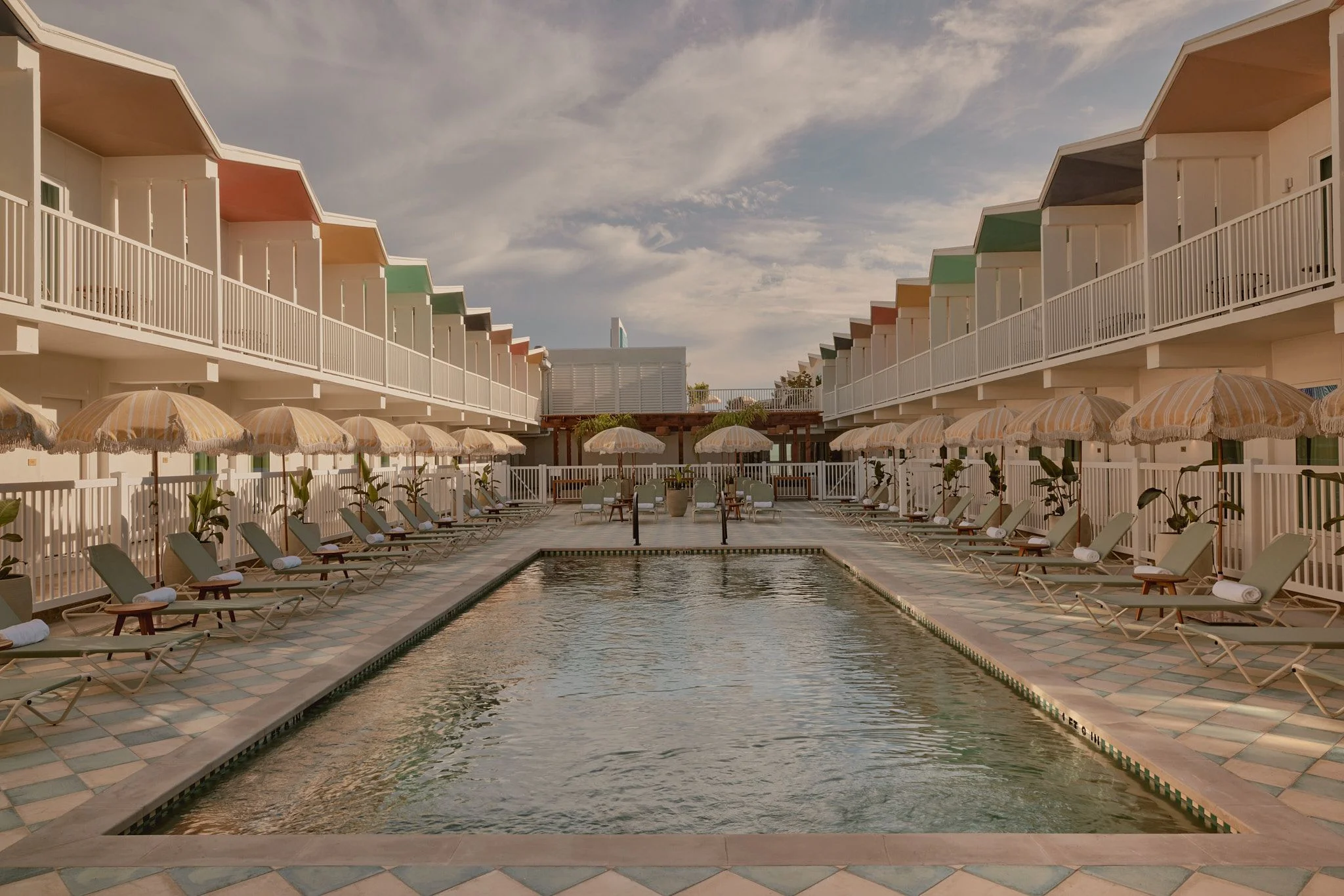 A symmetrical beachfront resort courtyard features a rectangular swimming pool flanked by rows of striped umbrellas and lounge chairs, with cream-colored beach houses featuring colorful roofs lining both sides and white railings under a soft, cloudy sky. The serene, nostalgic mid-century aesthetic is enhanced by manicured landscaping, terracotta paving, and a cohesive design palette of warm neutrals and vintage-inspired beach architecture.