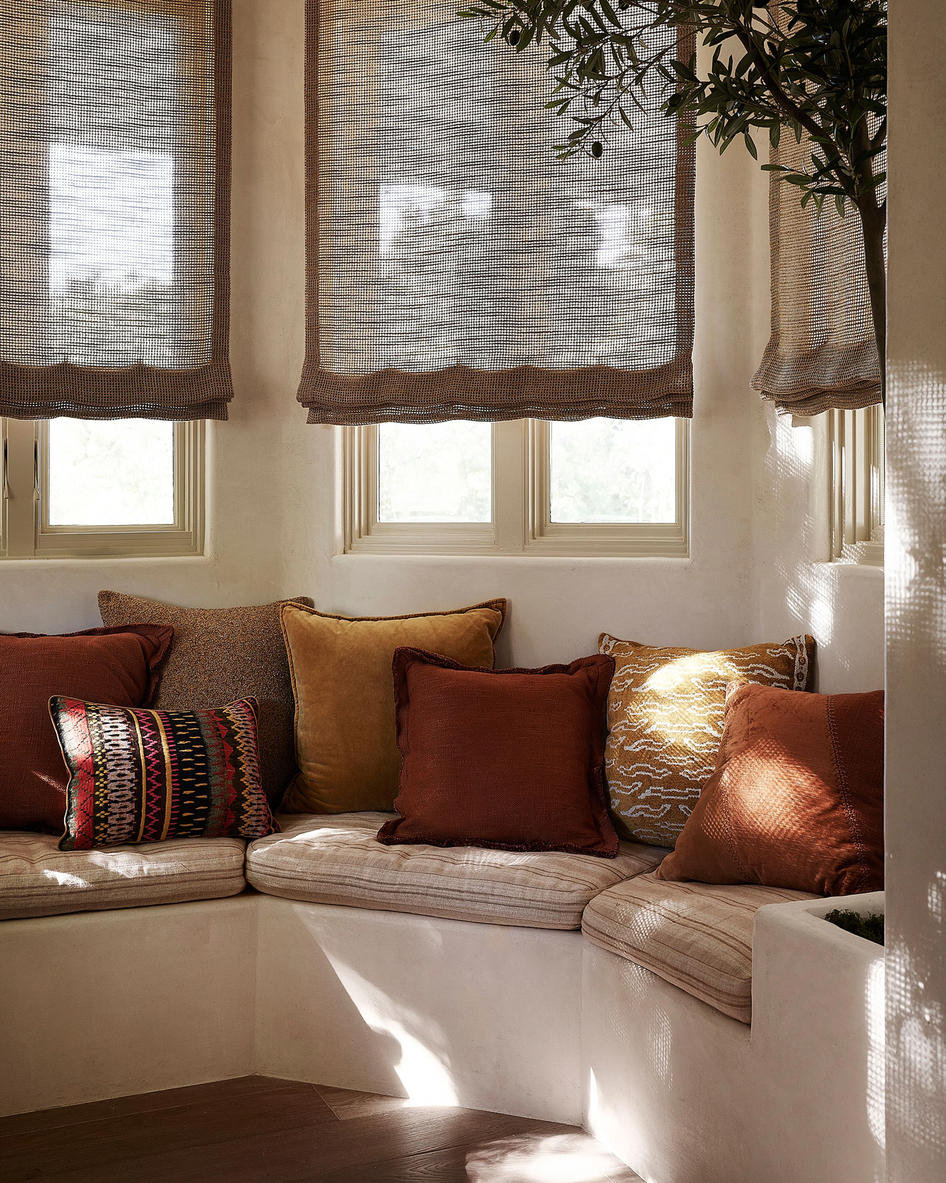 A bright, minimalist window nook features a built-in beige upholstered bench seat adorned with rust, ochre, and patterned decorative pillows, bathed in natural light filtered through three woven brown roman shades and cream-trimmed windows; a potted plant hangs from above, completing the warm, earthy aesthetic.