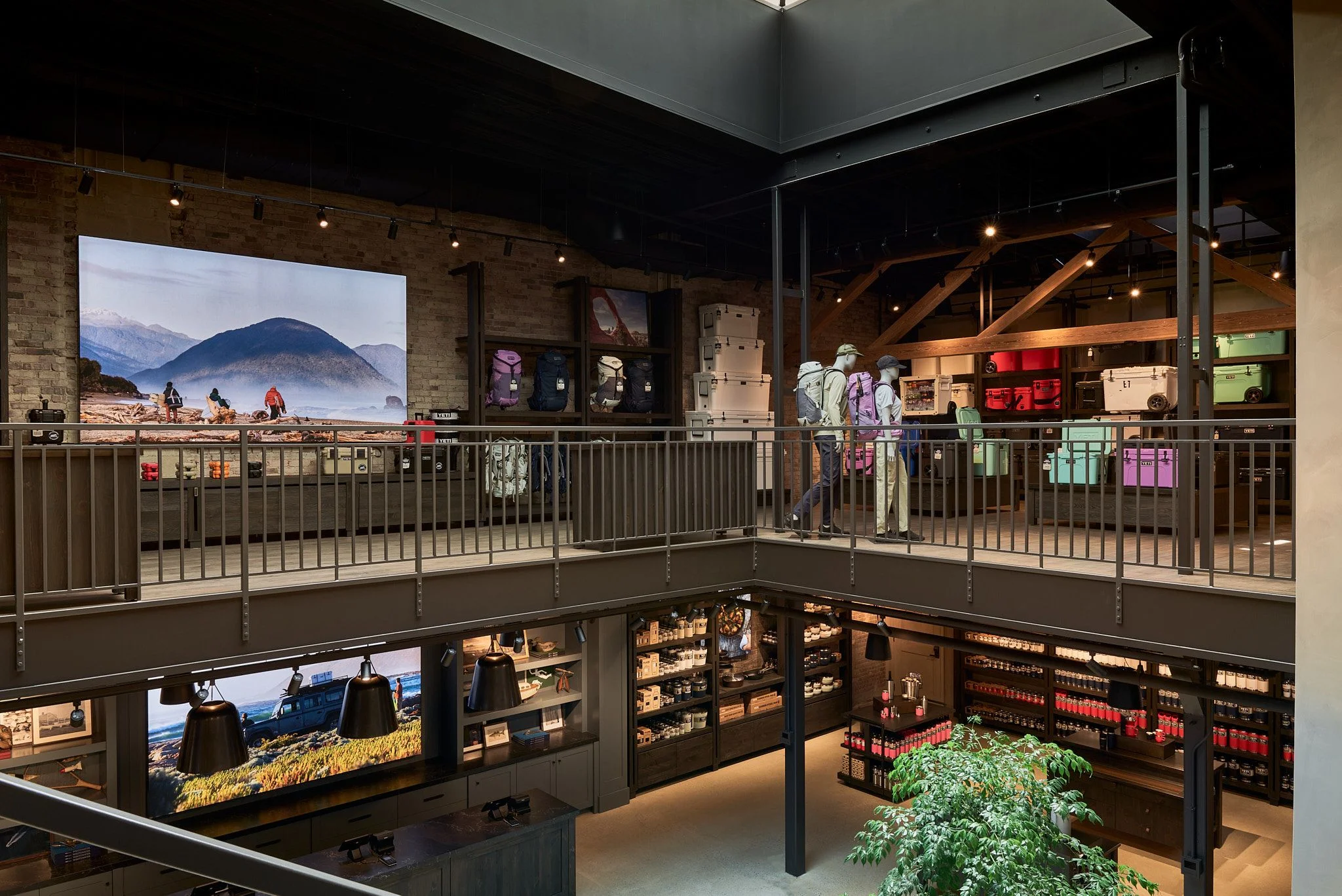 A modern retail showroom with an open two-level layout features exposed wooden beam ceiling, metal railings, and track lighting throughout. The upper mezzanine displays colorful hiking backpacks and outdoor gear on mannequins, while the lower level showcases illuminated display cases with products and large landscape photography.