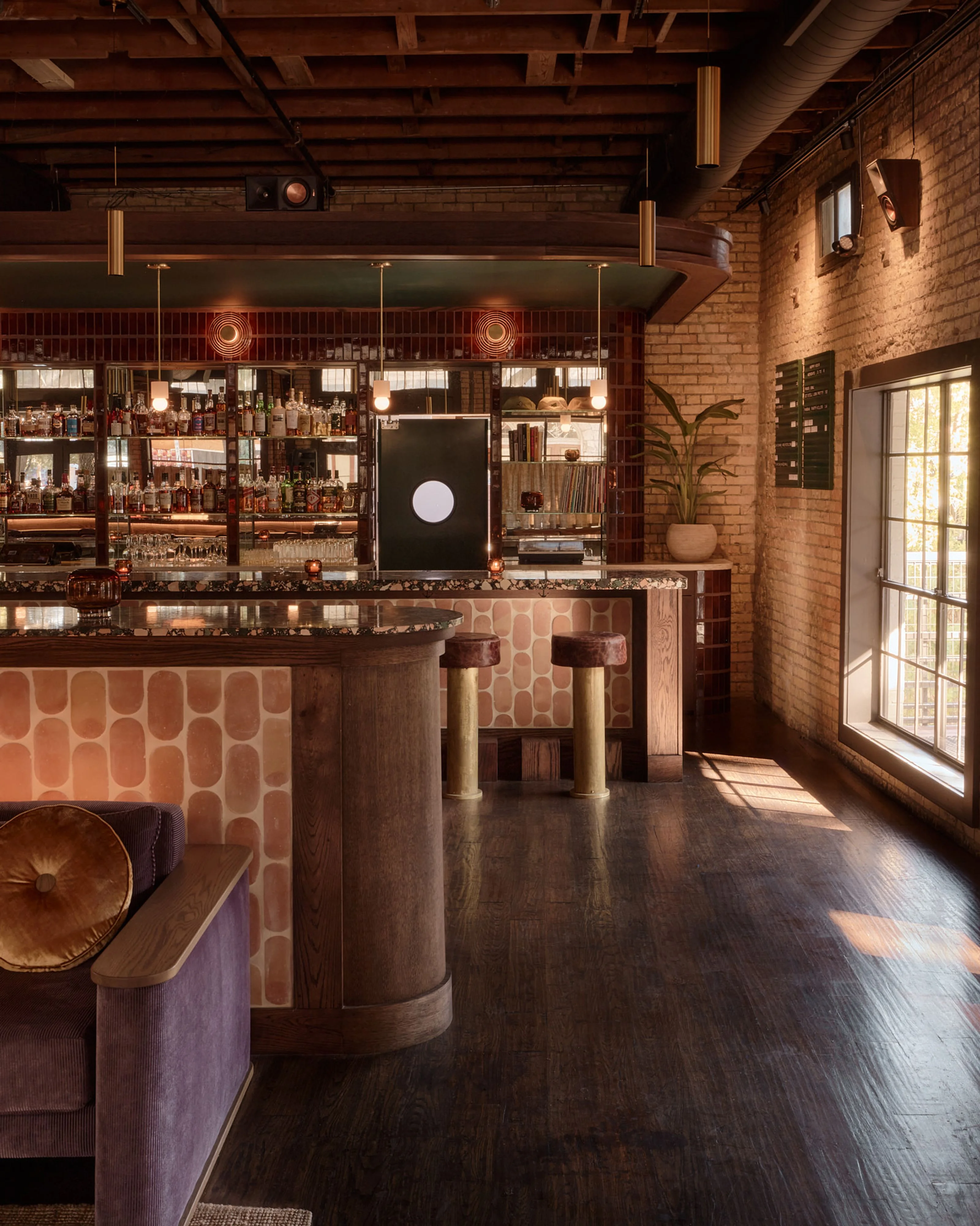 A warm, mid-century modern bar interior featuring exposed brick walls, wooden ceiling beams, and dark hardwood floors, with a prominent bar counter topped in terrazzo and accented with geometric patterned panels in rust and cream tones; vintage brass pendant lights and decorative wall sconces illuminate shelves stocked with bottles.