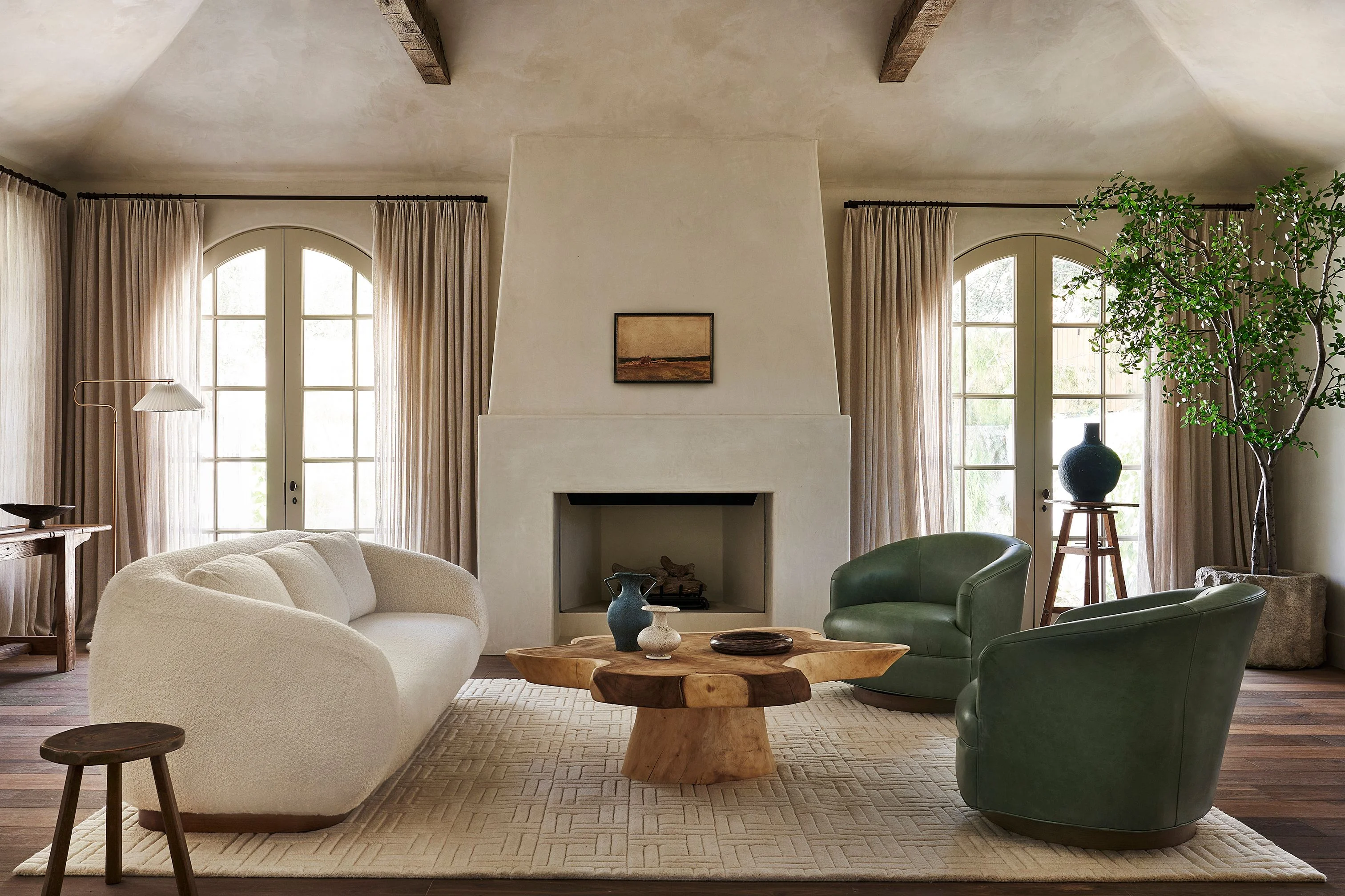 A serene, minimalist living room features a cream-colored curved sofa and two green leather armchairs arranged around a sculptural wooden coffee table, anchored by a neutral geometric rug. The space is unified by a prominent cream plaster fireplace with exposed wooden beams, tall arched windows with flowing linen curtains, and natural wood accents.