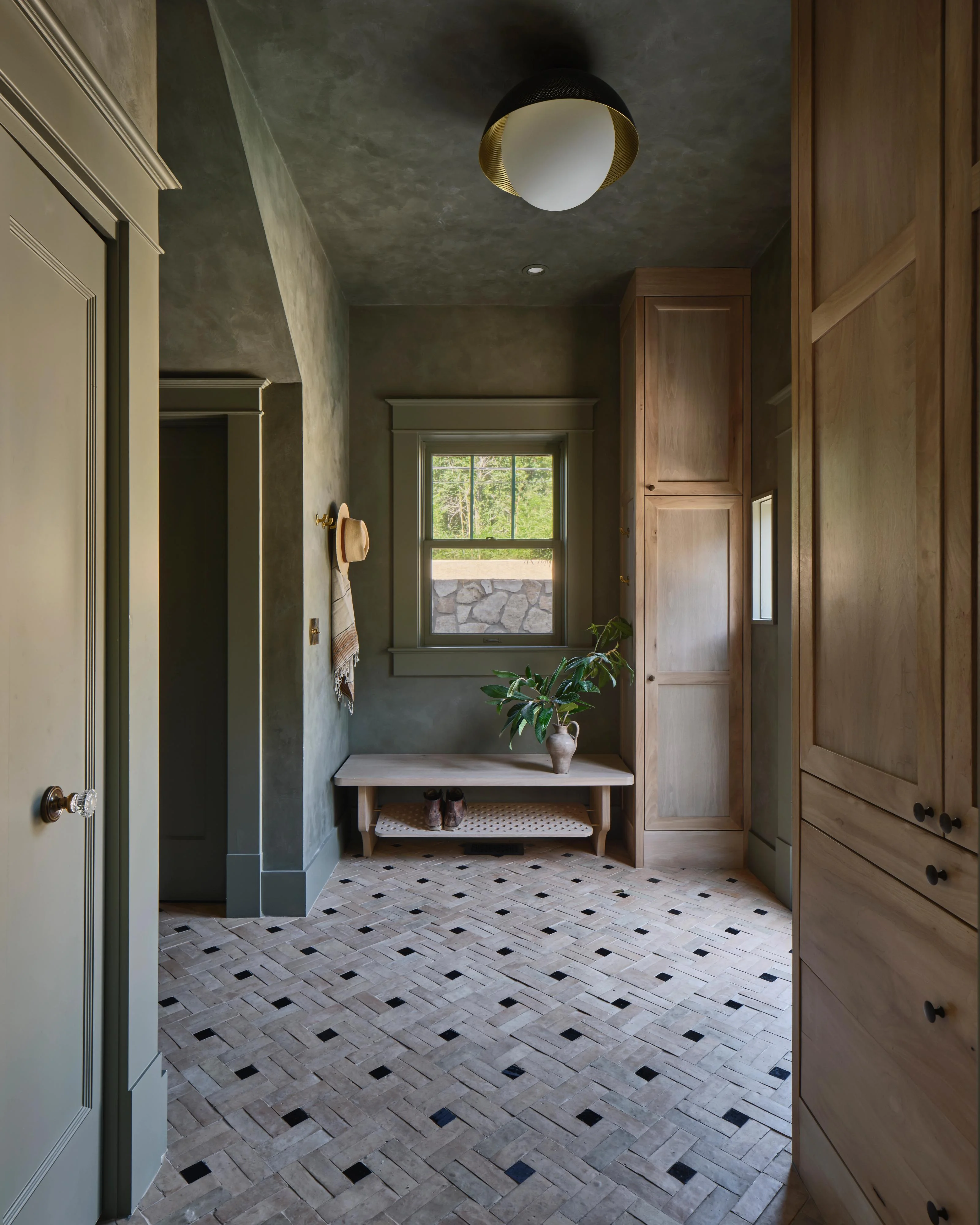 A sophisticated spa-like bathroom corridor features textured dark green walls, a geometric black-and-white herringbone tile floor, and natural wood cabinetry framing the space. A wooden bench with potted greenery sits beneath a window overlooking landscaping, while a brass-accented ceiling fixture and minimalist fixtures create a serene, contemporary aesthetic.