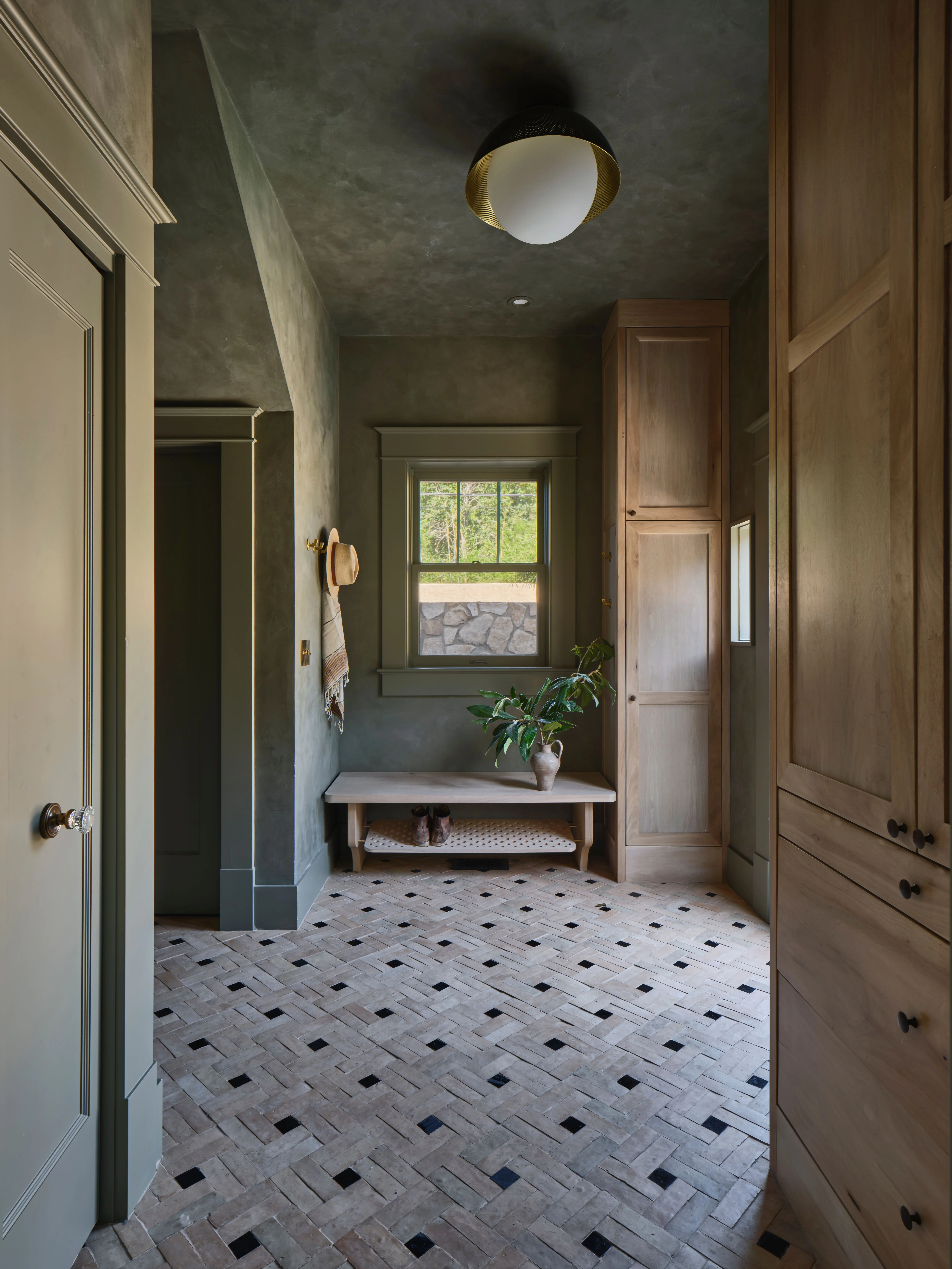 A sophisticated spa-like bathroom corridor features textured dark green walls, a geometric black-and-white herringbone tile floor, and natural wood cabinetry framing the space. A wooden bench with potted greenery sits beneath a window overlooking landscaping, while a brass-accented ceiling fixture and minimalist fixtures create a serene, contemporary aesthetic.