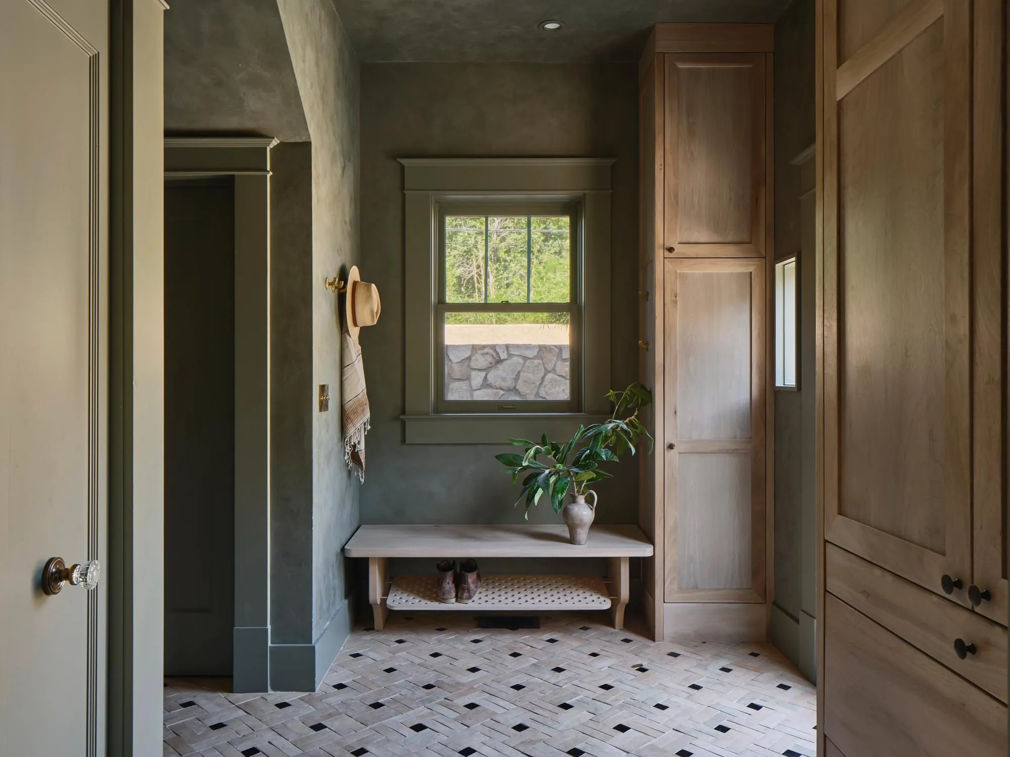 A sophisticated spa-like bathroom corridor features textured dark green walls, a geometric black-and-white herringbone tile floor, and natural wood cabinetry framing the space. A wooden bench with potted greenery sits beneath a window overlooking landscaping, while a brass-accented ceiling fixture and minimalist fixtures create a serene, contemporary aesthetic.