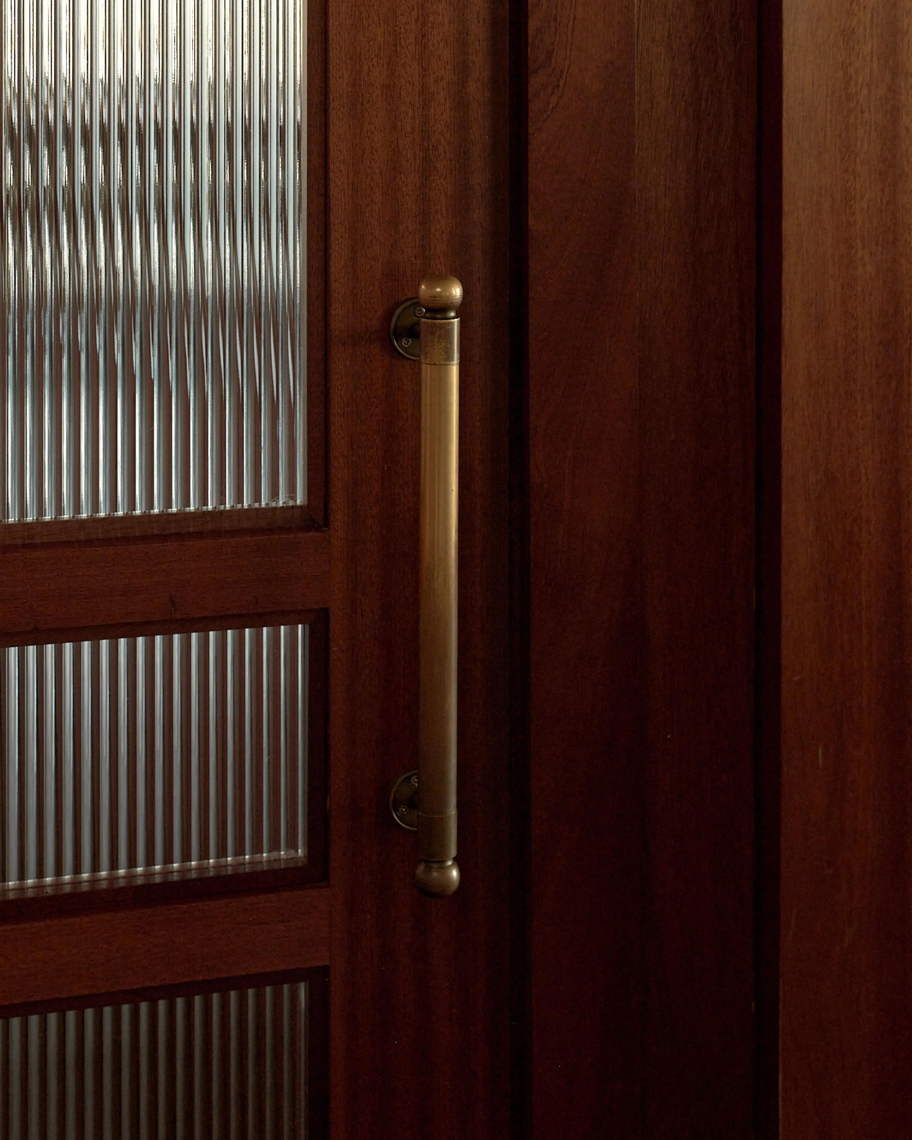 A minimalist architectural detail showing a rich brown wooden door with three horizontal panels of reeded glass, accented by a long brass cylindrical handle with spherical finials. The warm wood tones and industrial metal hardware create a striking mid-century modern aesthetic.