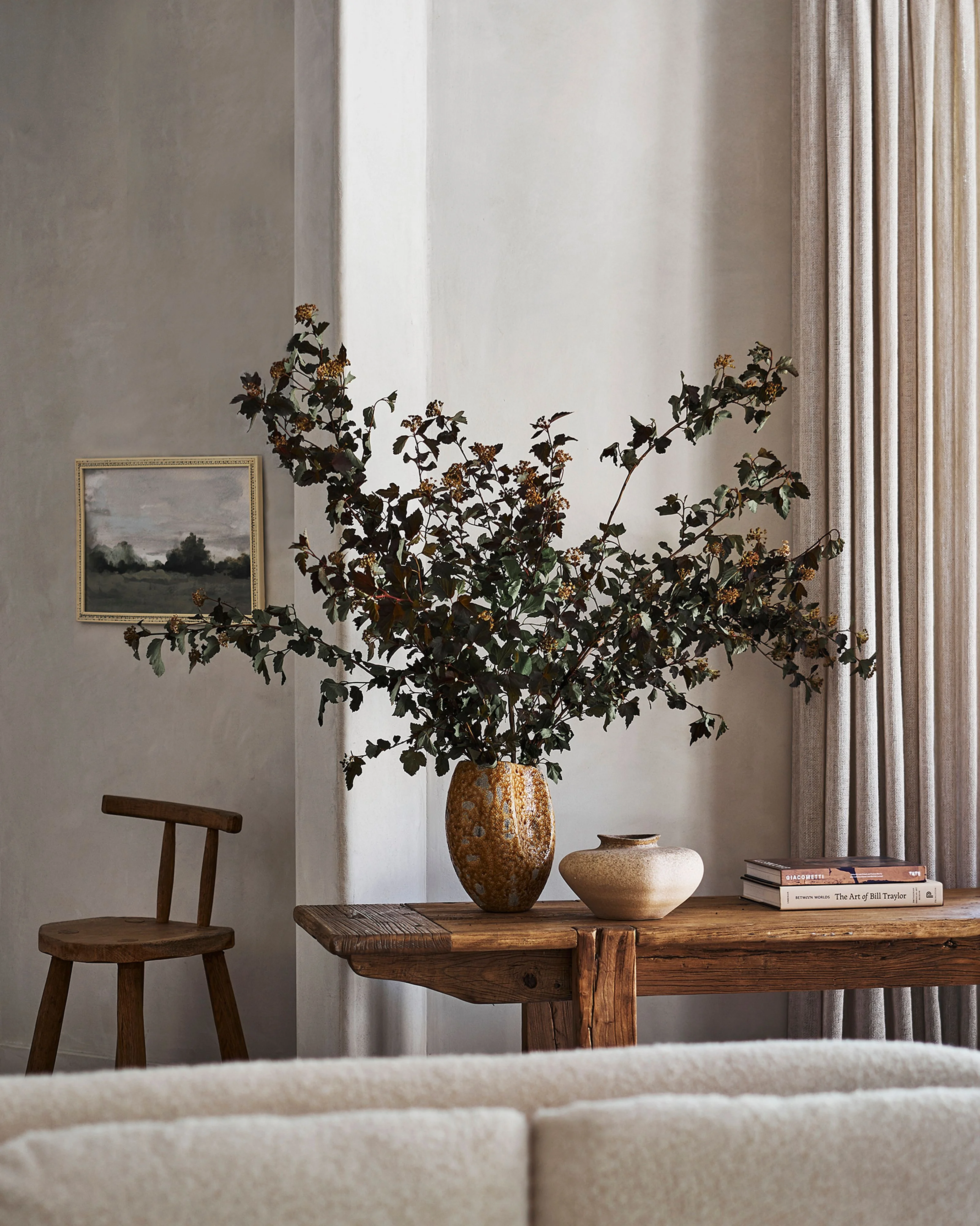 A minimalist, light-filled interior featuring a rustic wooden table adorned with dried foliage in textured ceramic vessels, a simple wooden chair, and stacked books, set against neutral beige walls, cream curtains, and a framed landscape painting.
