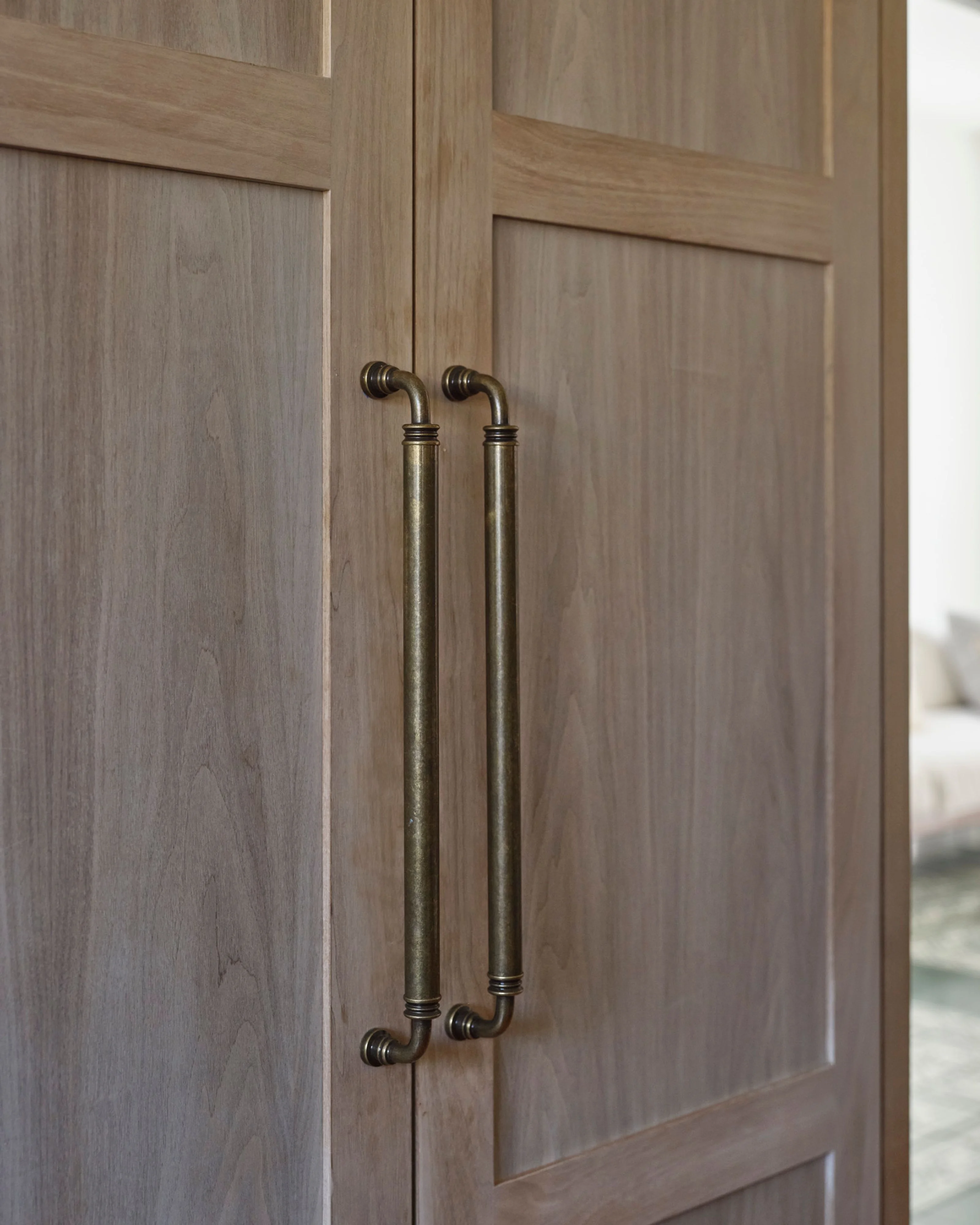 A close-up of a pair of light gray-stained wooden cabinet doors featuring raised panel detailing, fitted with elegant brass curved handles with decorative finials.