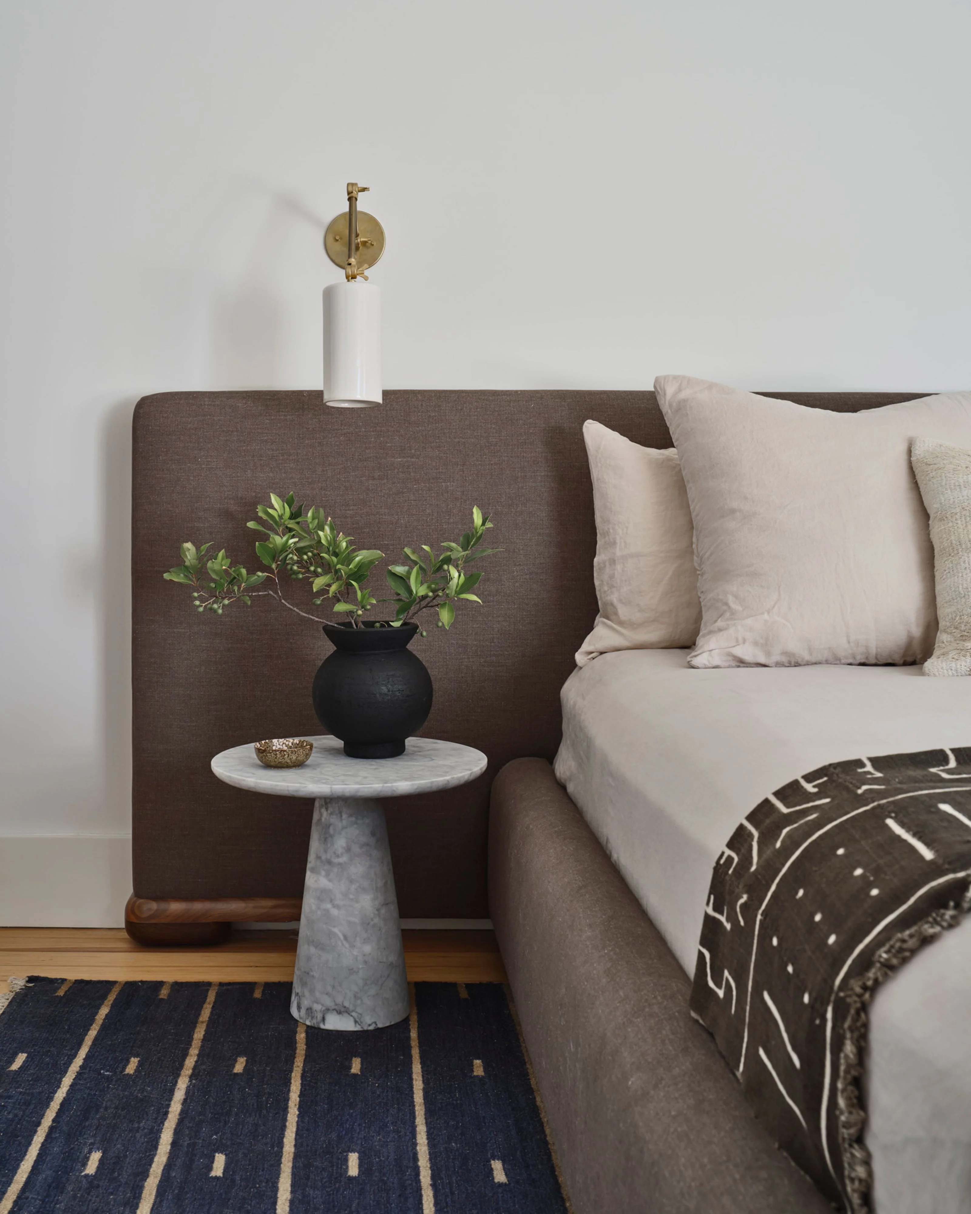 A serene bedroom corner featuring a charcoal-brown upholstered headboard with a white wall behind it, accessorized with a brass wall sconce. A gray concrete pedestal side table holds a black spherical vase with green foliage, positioned next to a bed dressed in cream linens and a geometric patterned throw blanket.