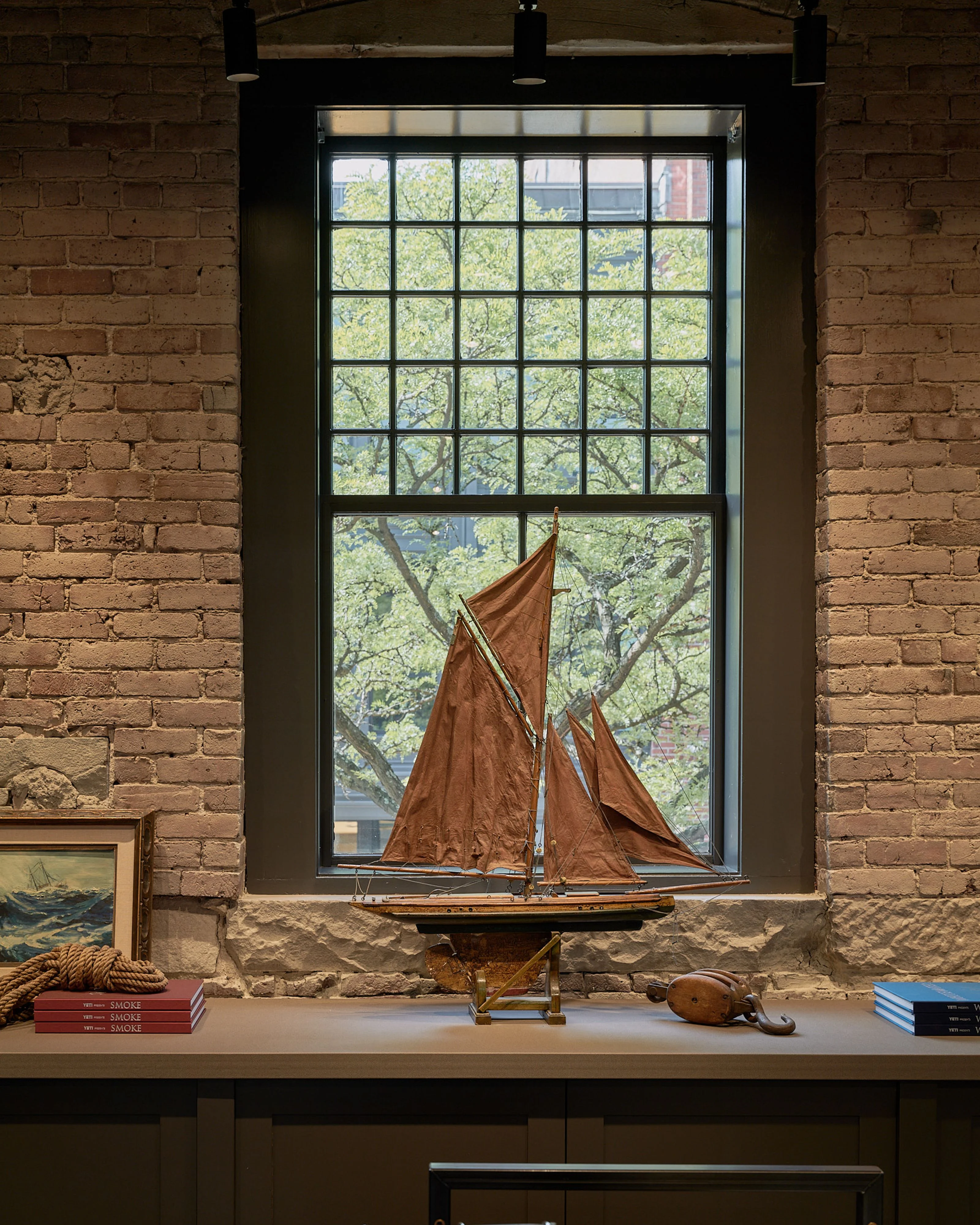 A rustic brick interior features a large industrial-style window with a black frame and gridded panes, framing leafy green trees outside. On the stone windowsill below sits a wooden sailing ship model with rust-colored fabric sails, accompanied by nautical decor including stacked books, rope, and wooden objects.