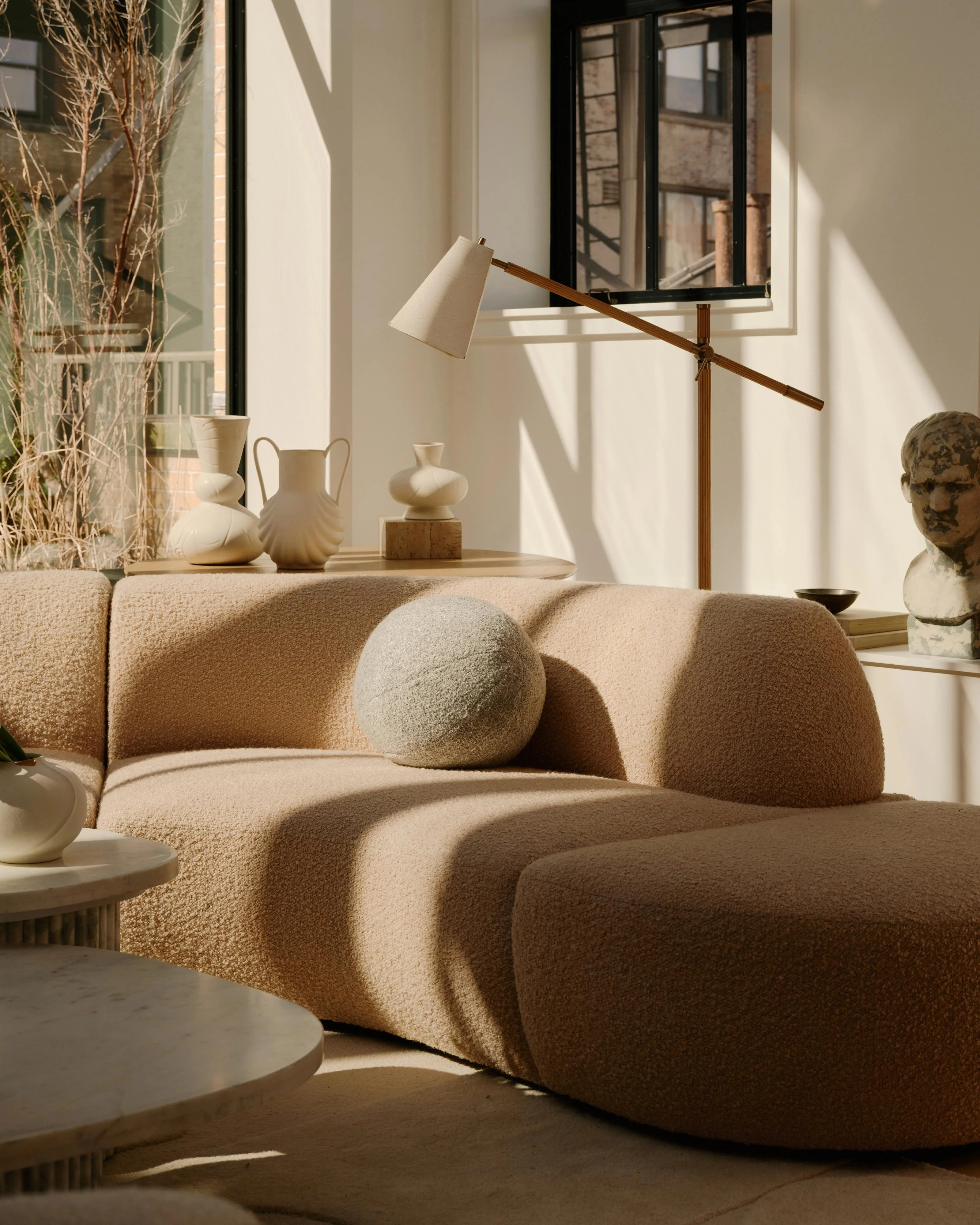 A minimalist living room with a sculptural curved beige sofa featuring organic, rounded forms and textured upholstery, accented by a spherical throw pillow and complemented by neutral ceramic vessels, a wooden-armed floor lamp, and a classical bust, all bathed in natural light from black-framed windows.