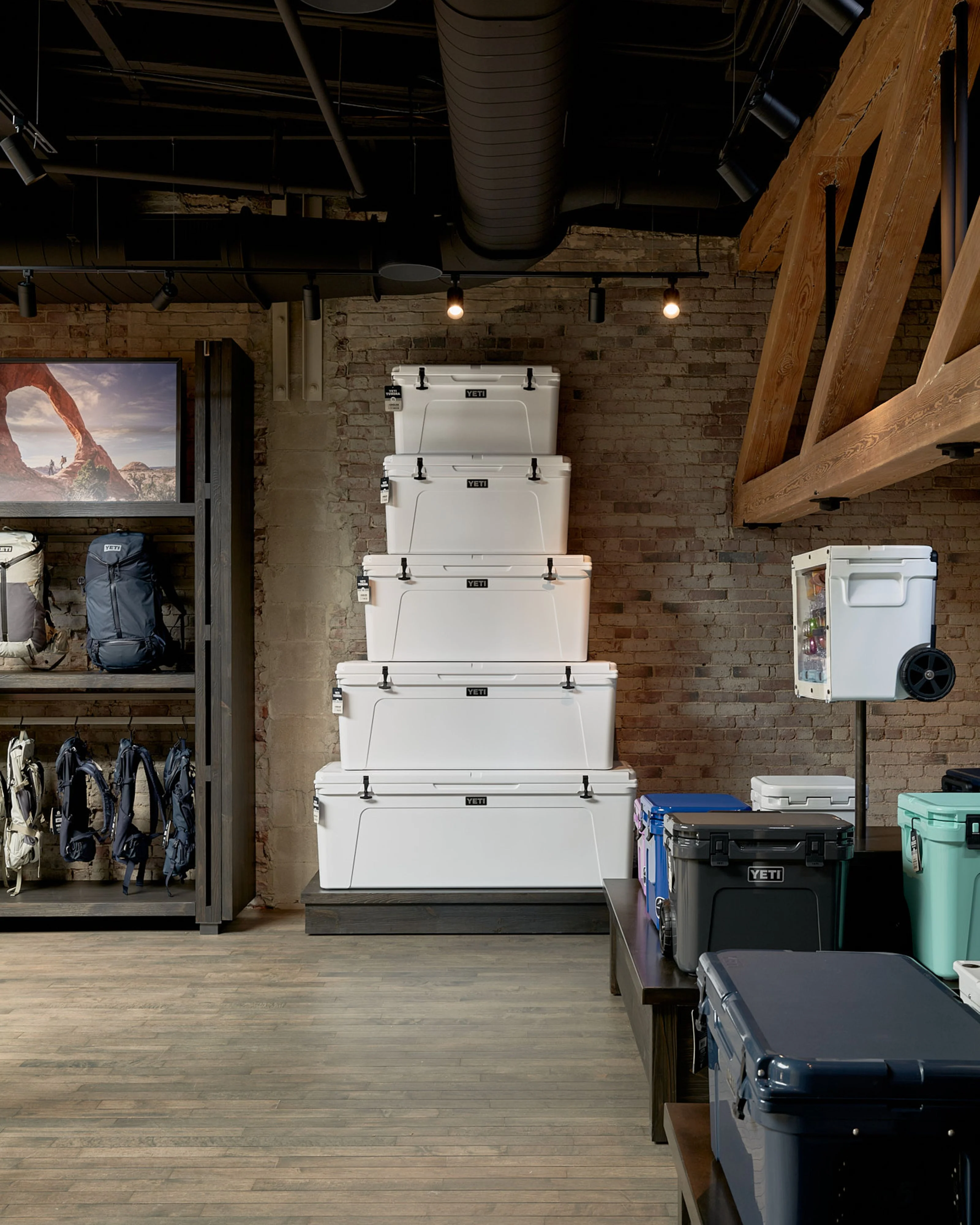 An industrial-style retail showroom features an exposed brick wall with stacked white coolers as a central display, complemented by raw wooden beams, exposed ductwork, and track lighting overhead. The space showcases outdoor gear and equipment on shelving and displays, with wooden flooring and a modern, minimalist aesthetic.