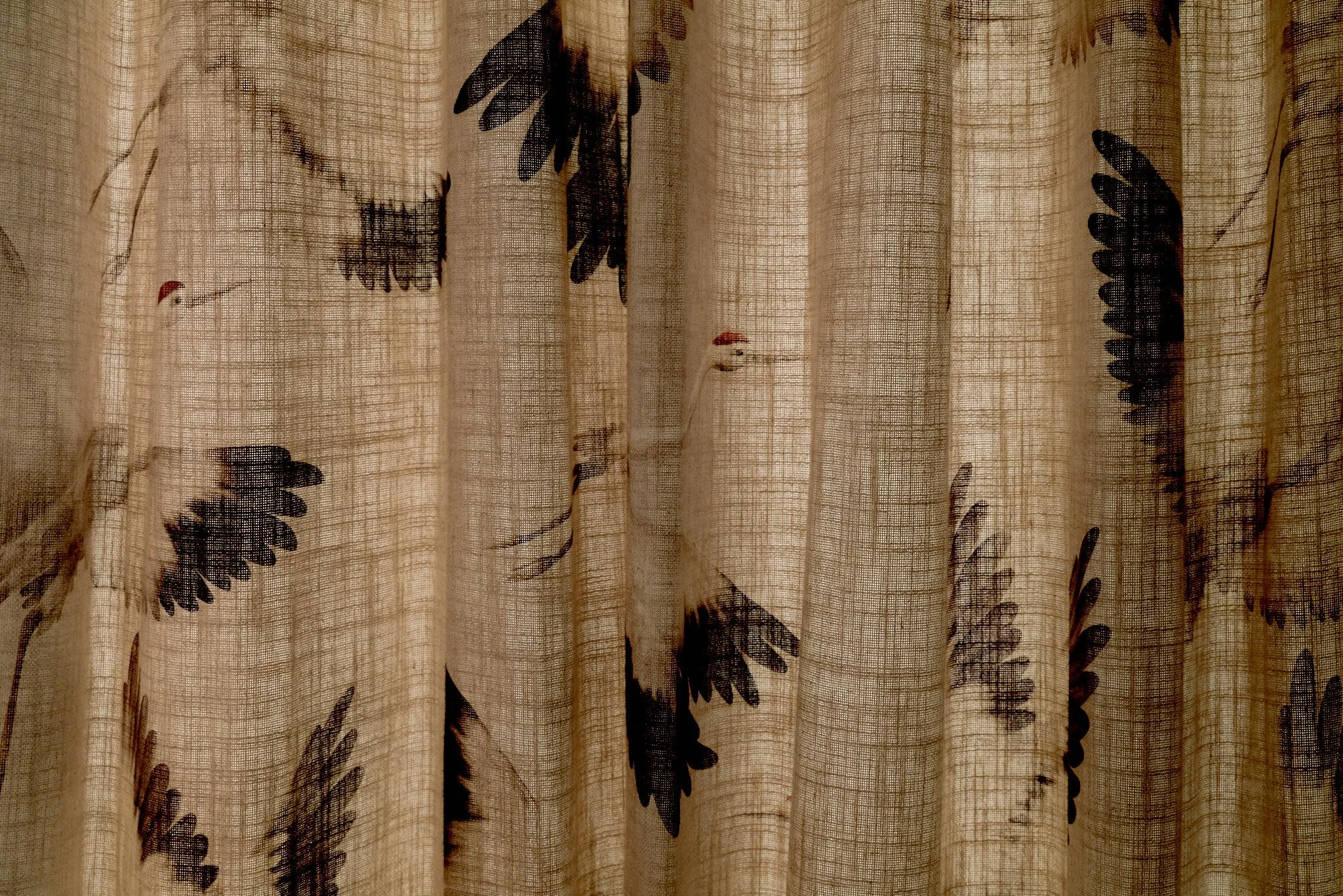 Sheer linen curtains with a botanical feather motif in black and burgundy hang in vertical pleats, creating a warm, textured interior with soft diffused light filtering through the natural fabric.