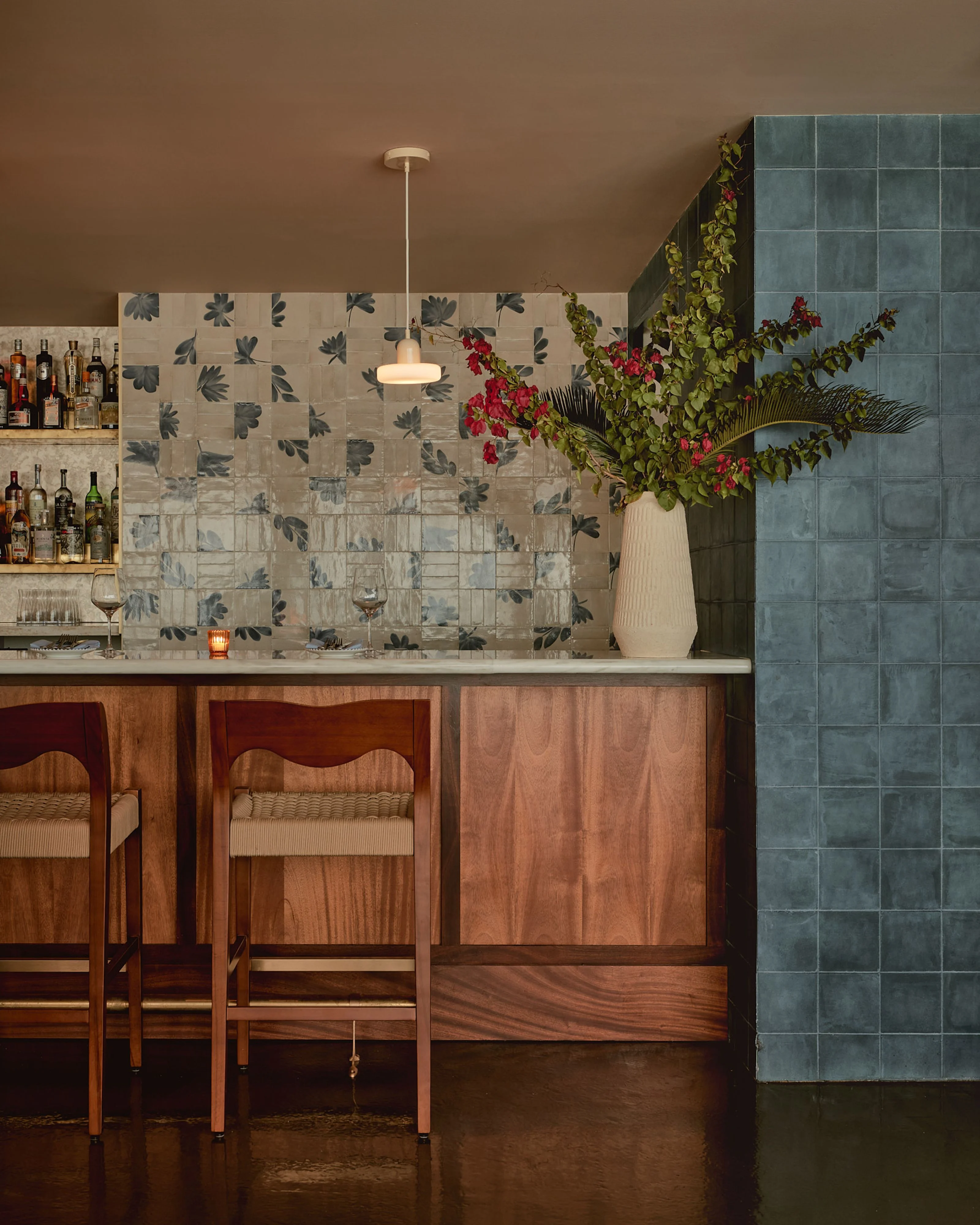 A warm, sophisticated bar interior featuring warm brown walls and a wooden counter with mid-century modern bar stools, accented by a decorative leaf-patterned tile backsplash, a teal-blue tiled wall, and a large cream vase with cascading red flowers and greenery serving as a striking botanical centerpiece.