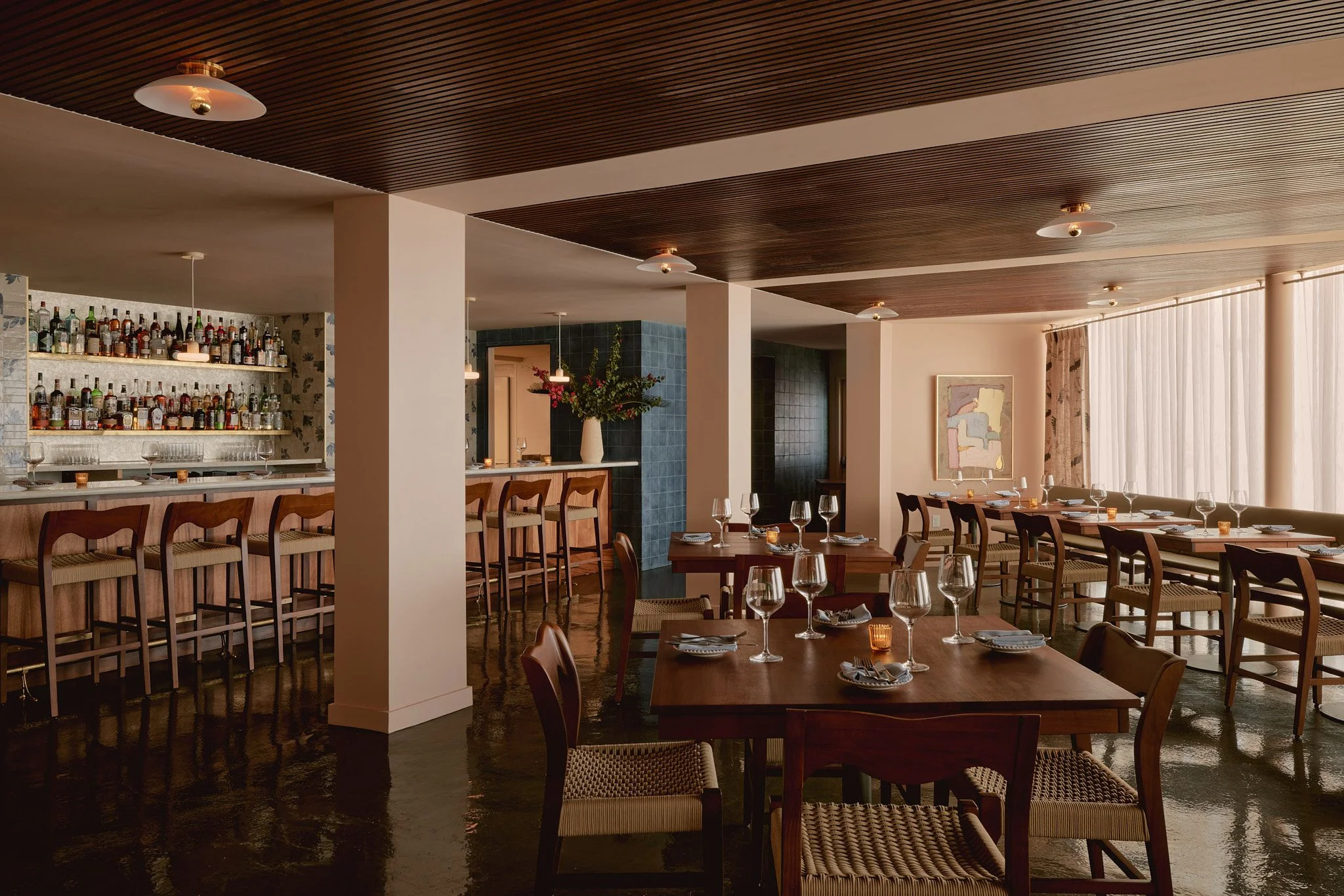 A modern mid-century restaurant interior features warm wood tones, polished dark floors, and a coffered ceiling with recessed lighting. The open-concept space includes a well-stocked bar with bottle displays on the left, dining tables set with glassware in the center, and floor-to-ceiling windows on the right, creating an elegant atmosphere enhanced by natural light and contemporary wood furnishings.