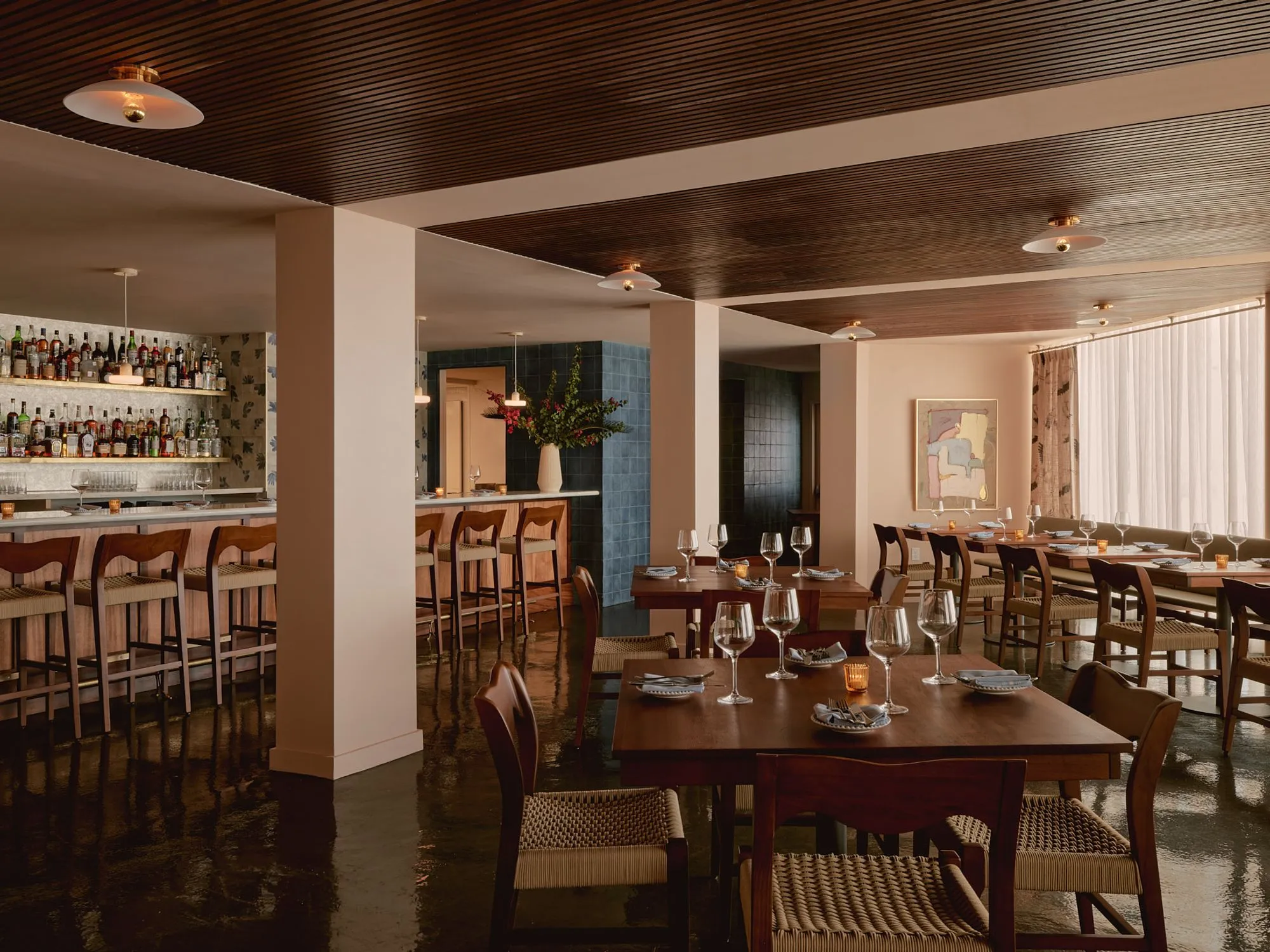 A modern mid-century restaurant interior features warm wood tones, polished dark floors, and a coffered ceiling with recessed lighting. The open-concept space includes a well-stocked bar with bottle displays on the left, dining tables set with glassware in the center, and floor-to-ceiling windows on the right, creating an elegant atmosphere enhanced by natural light and contemporary wood furnishings.