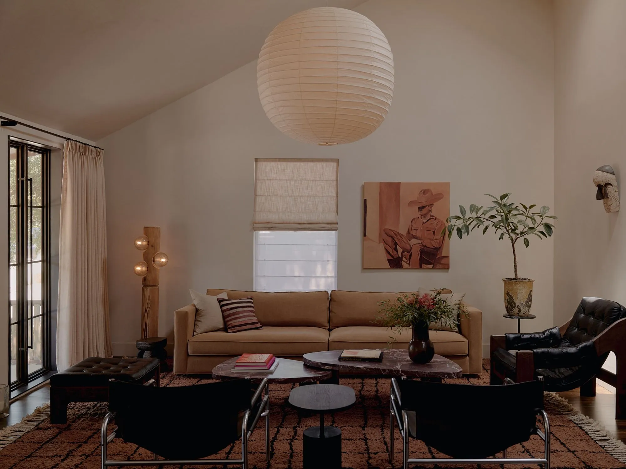 A modern living room with warm, neutral tones features a tan upholstered sofa beneath a large white paper pendant lamp, anchored by a terracotta-patterned area rug and complemented by black leather lounge chairs, potted plants, and warm wood accents throughout the minimalist space.