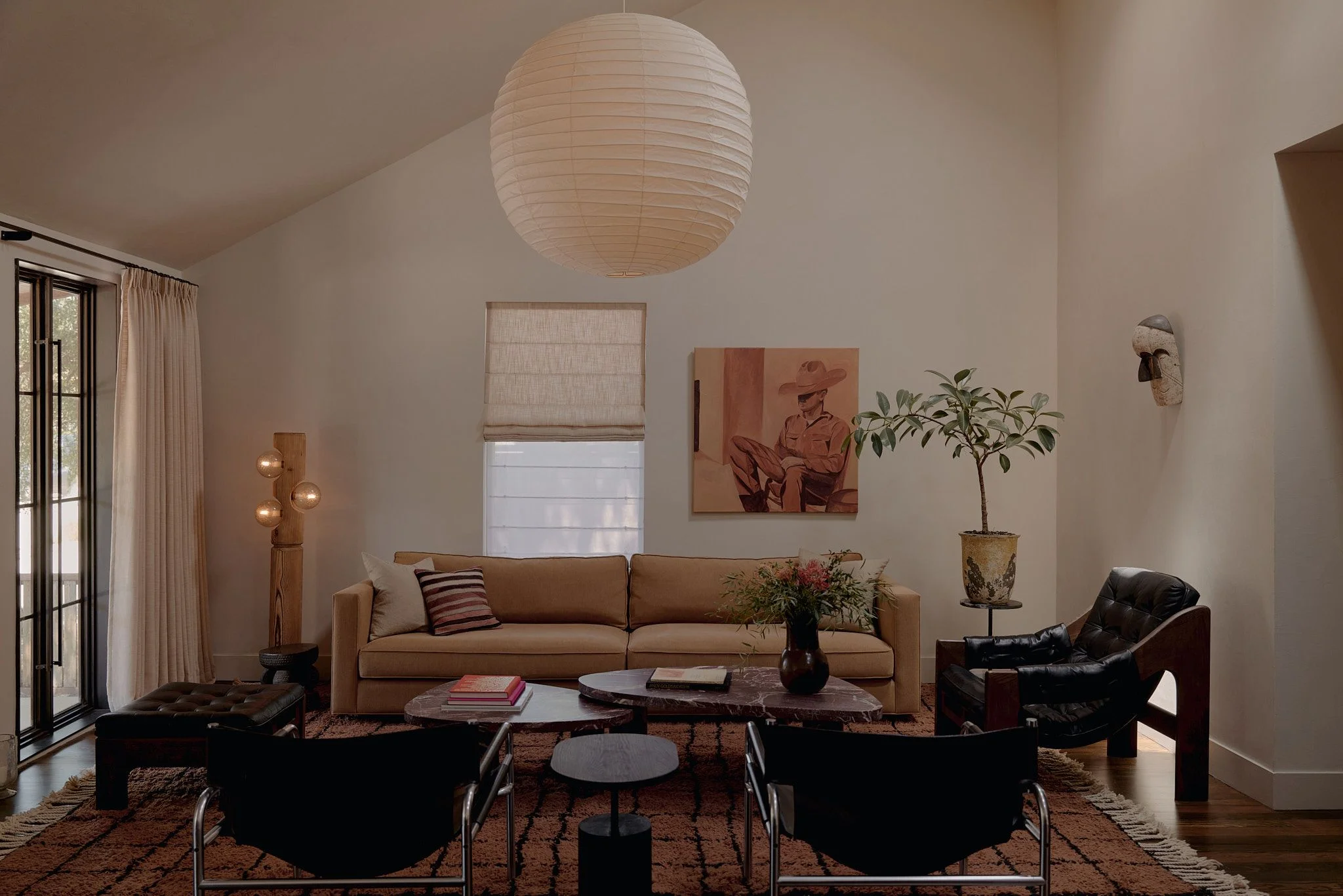 A modern living room with warm, neutral tones features a tan upholstered sofa beneath a large white paper pendant lamp, anchored by a terracotta-patterned area rug and complemented by black leather lounge chairs, potted plants, and warm wood accents throughout the minimalist space.
