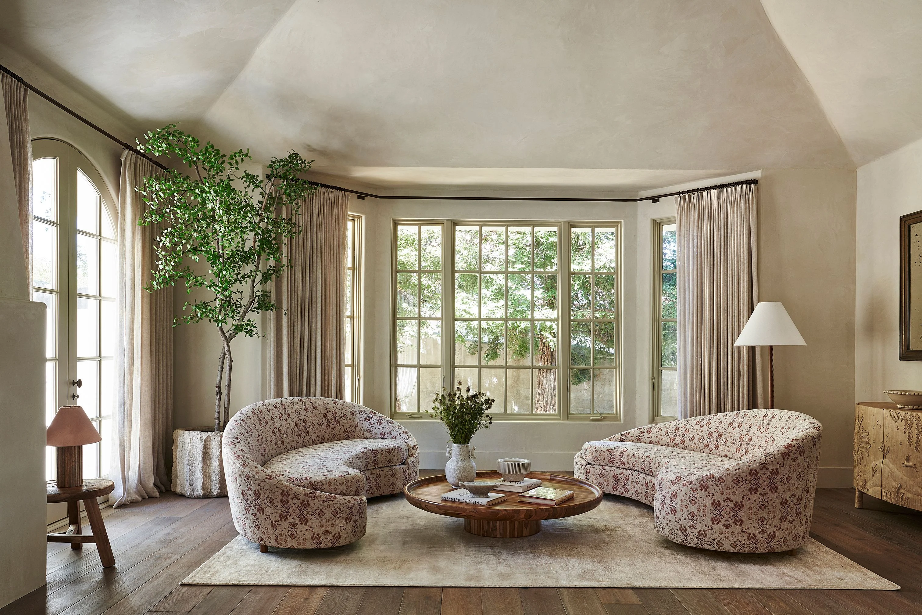 A serene, light-filled living room with curved cream-upholstered chairs arranged around a wooden circular table, featuring expansive multi-pane windows framed by floor-to-ceiling neutral curtains, vaulted ceilings, and natural elements including potted greenery and a minimalist botanical accent piece.